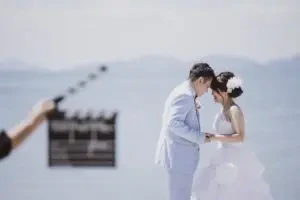 玉島の海を背景に、カチンコを構えたスタッフの前で新郎新婦様が顔を寄せ合う撮影中のワンシーンです