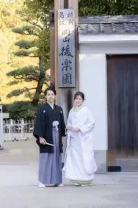 岡山後楽園の入り口前で紋付袴の新郎様と白無垢姿の新婦様が並んで立つ様子
