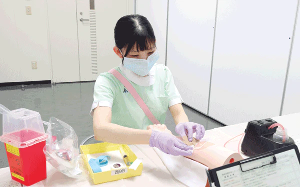 IVナース認定試験では、血管の選択に関する口頭試問の後、ベッドサイドでの採血の流れを再現するかたちで実施される。患者さんへの声かけからフルネームの確認、手技、痛みや腫れの確認、廃棄物の始末、退出までが