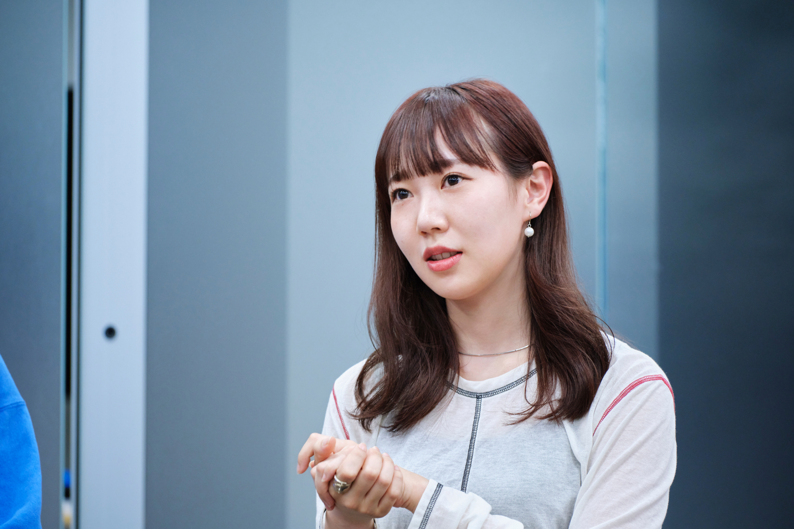 長い茶色の若い女性。白いトップスを着て、真剣な表情で何かを説明しているように見える。耳にはシンプルなピアスをつけている。