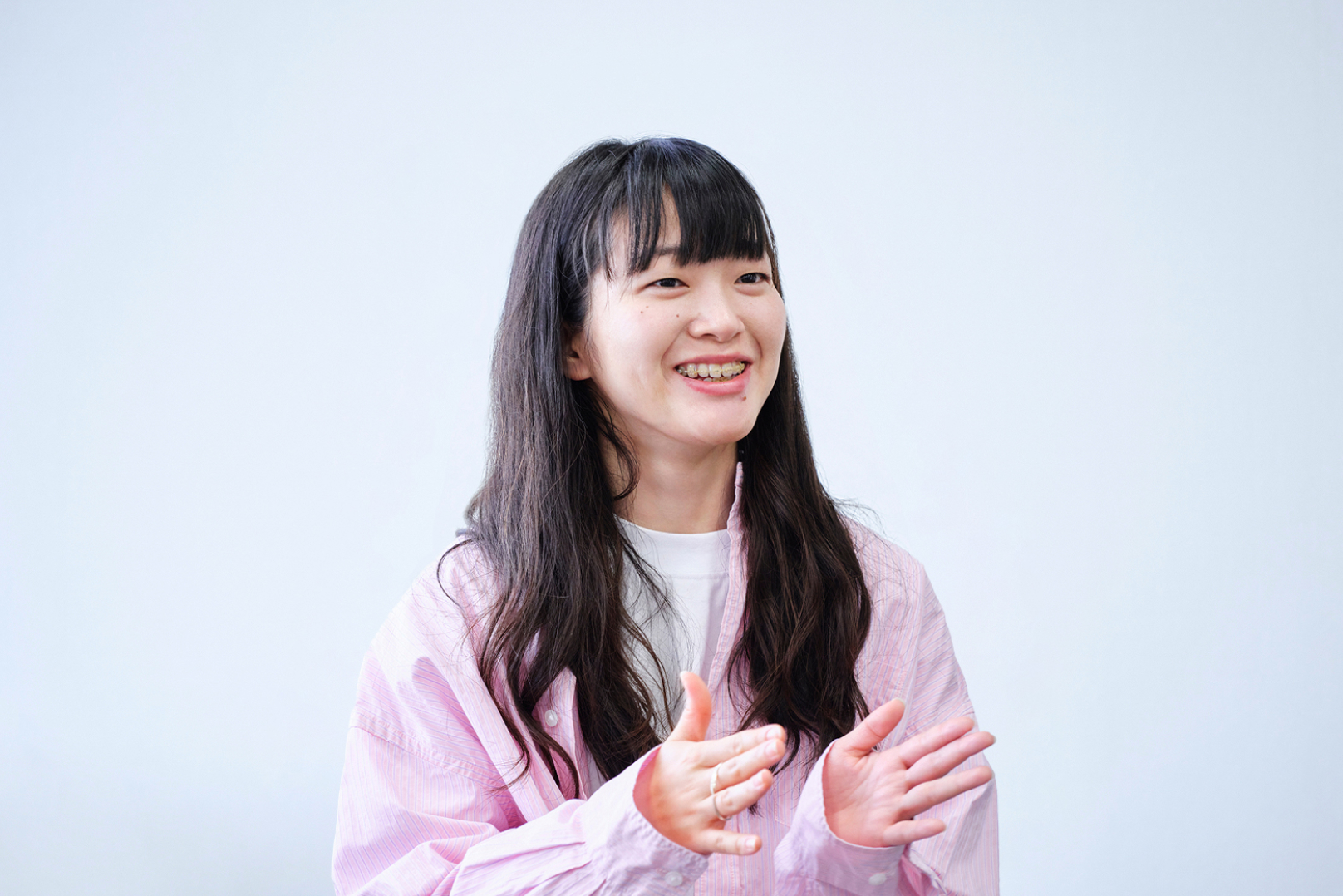 笑顔を見せる、長い黒髪の女性。ピンク色のシャツを着ている。