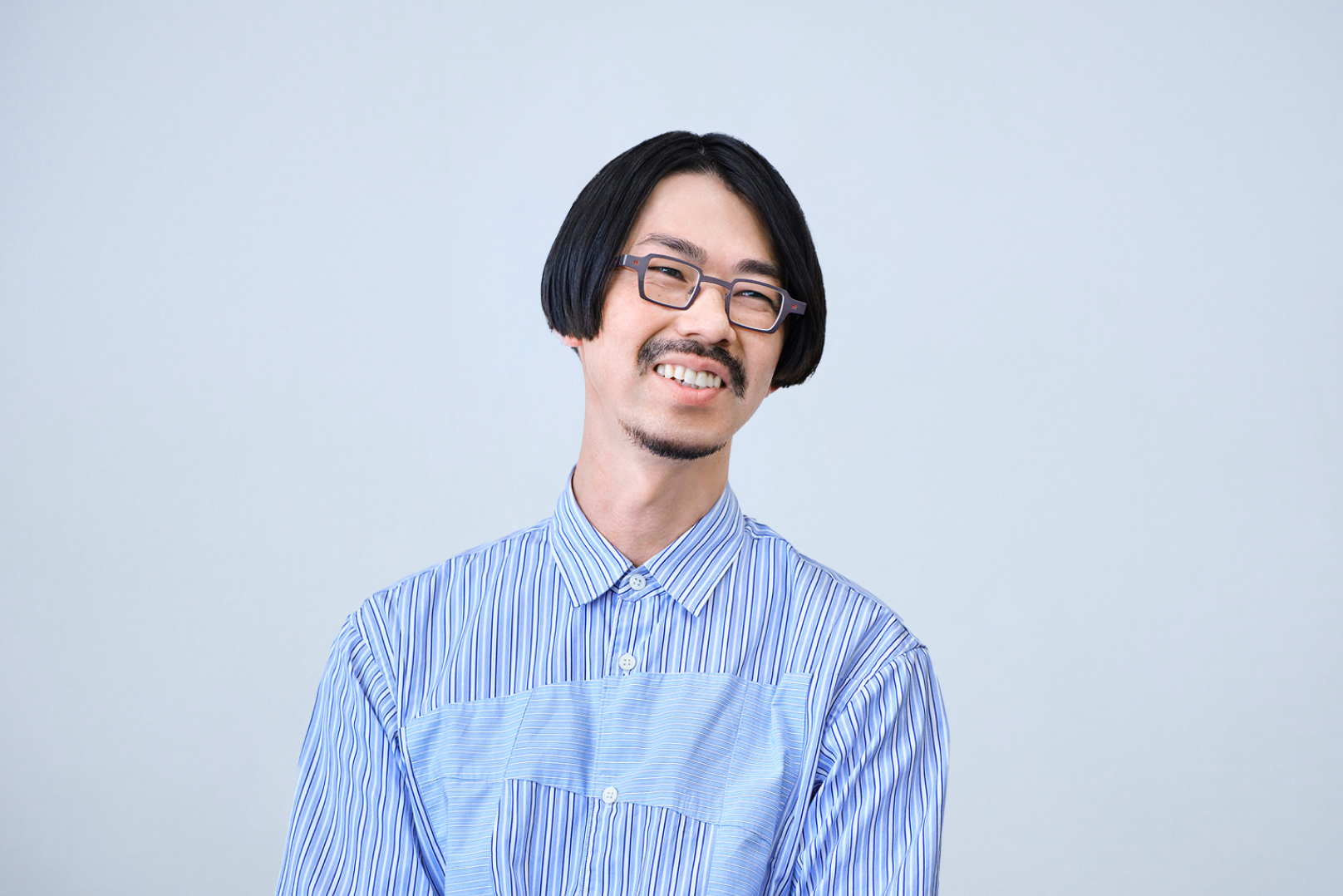 青いストライプのシャツを着た、眼鏡をかけた笑顔の男性の上半身写真。