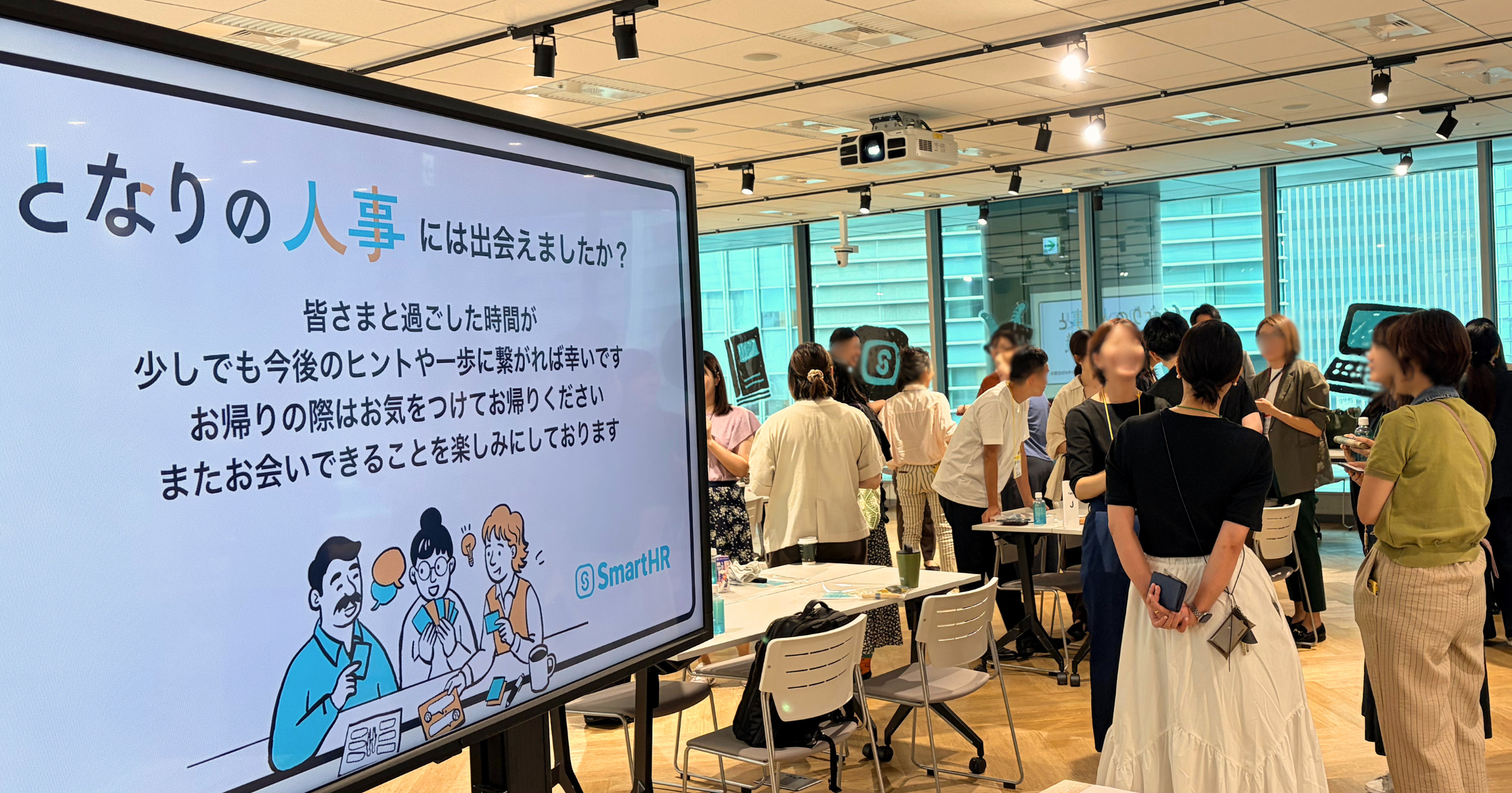 「となりの人事には出会えましたか？」と表示されたモニターと、会場内で立ち話をする人々の様子