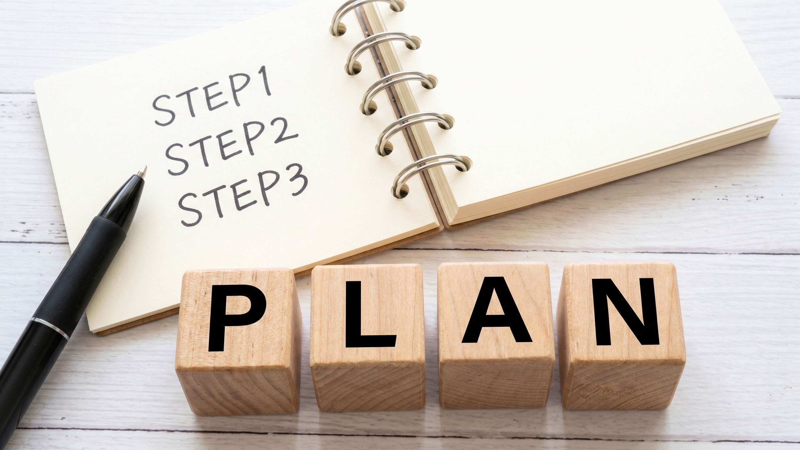 「STEP1 STEP2 STEP3」と書かれたノートと黒いペン、その手前に「PLAN」の文字が並ぶ木製ブロック
