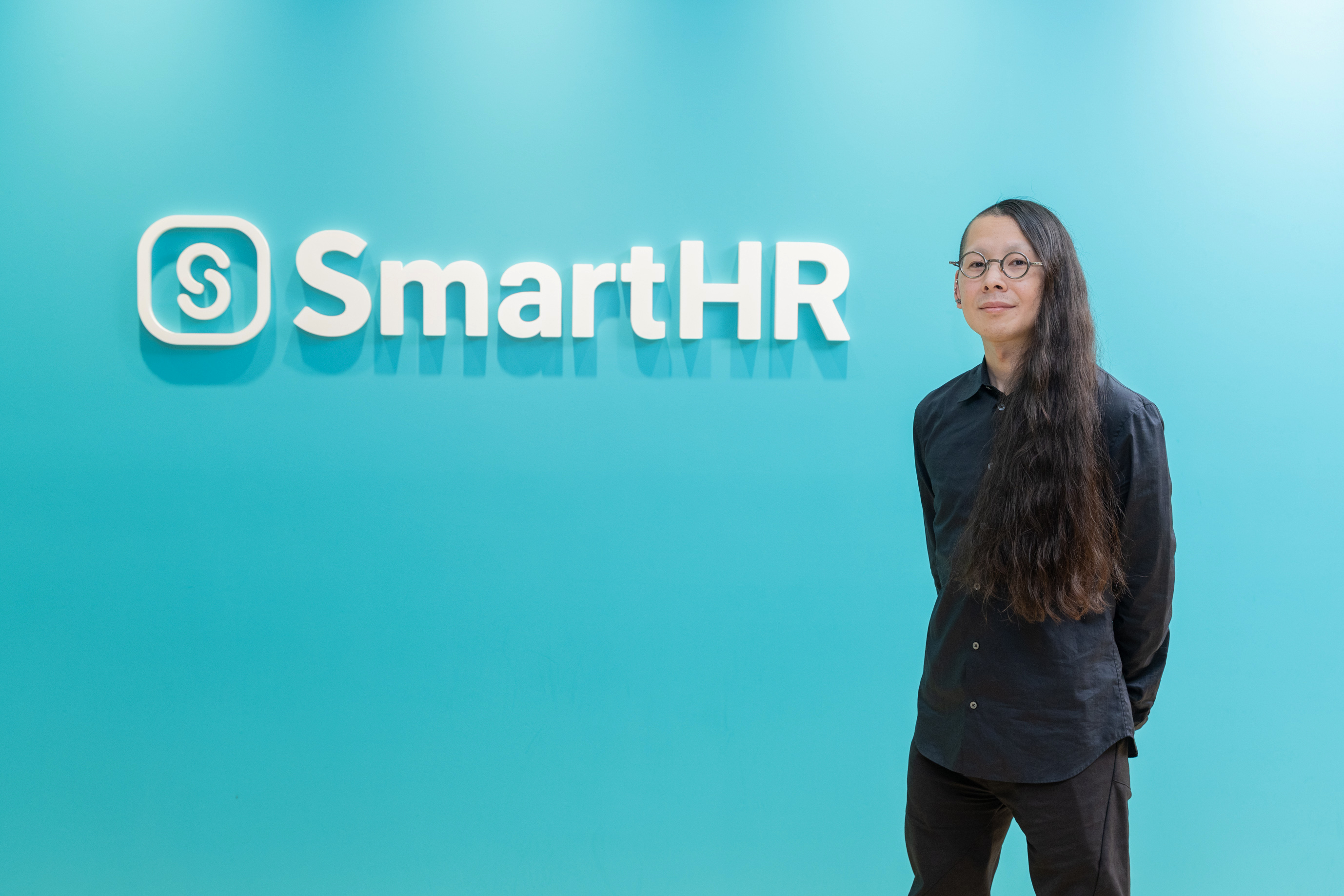 SmartHRのロゴが配置された青緑色の壁の前に立つ、黒いシャツ姿のロングヘアの男性（宮原）。