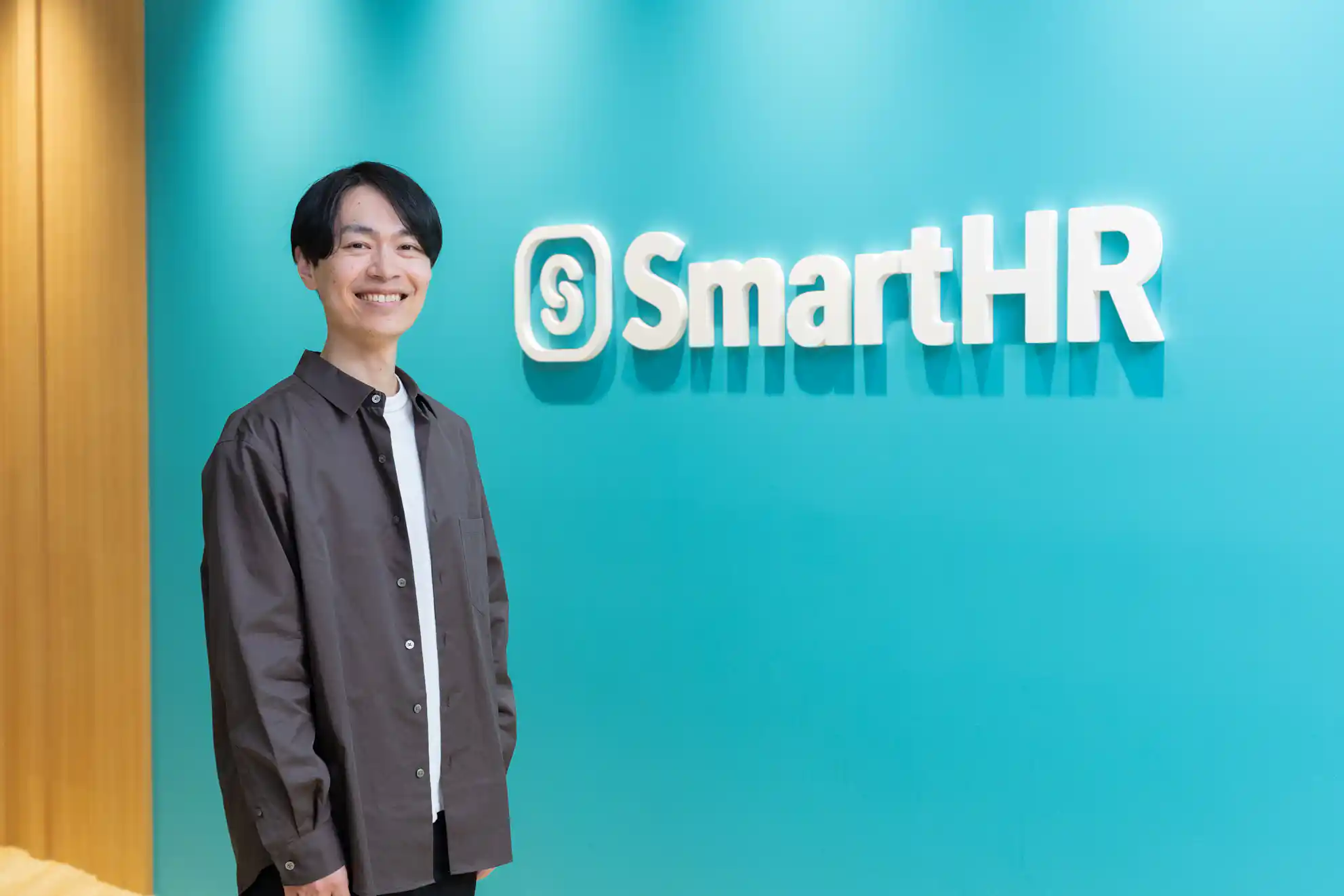 「SmartHR」の白いロゴが掲げられた水色の壁の前に立ち、笑顔で正面を見ている茶色いシャツの男性