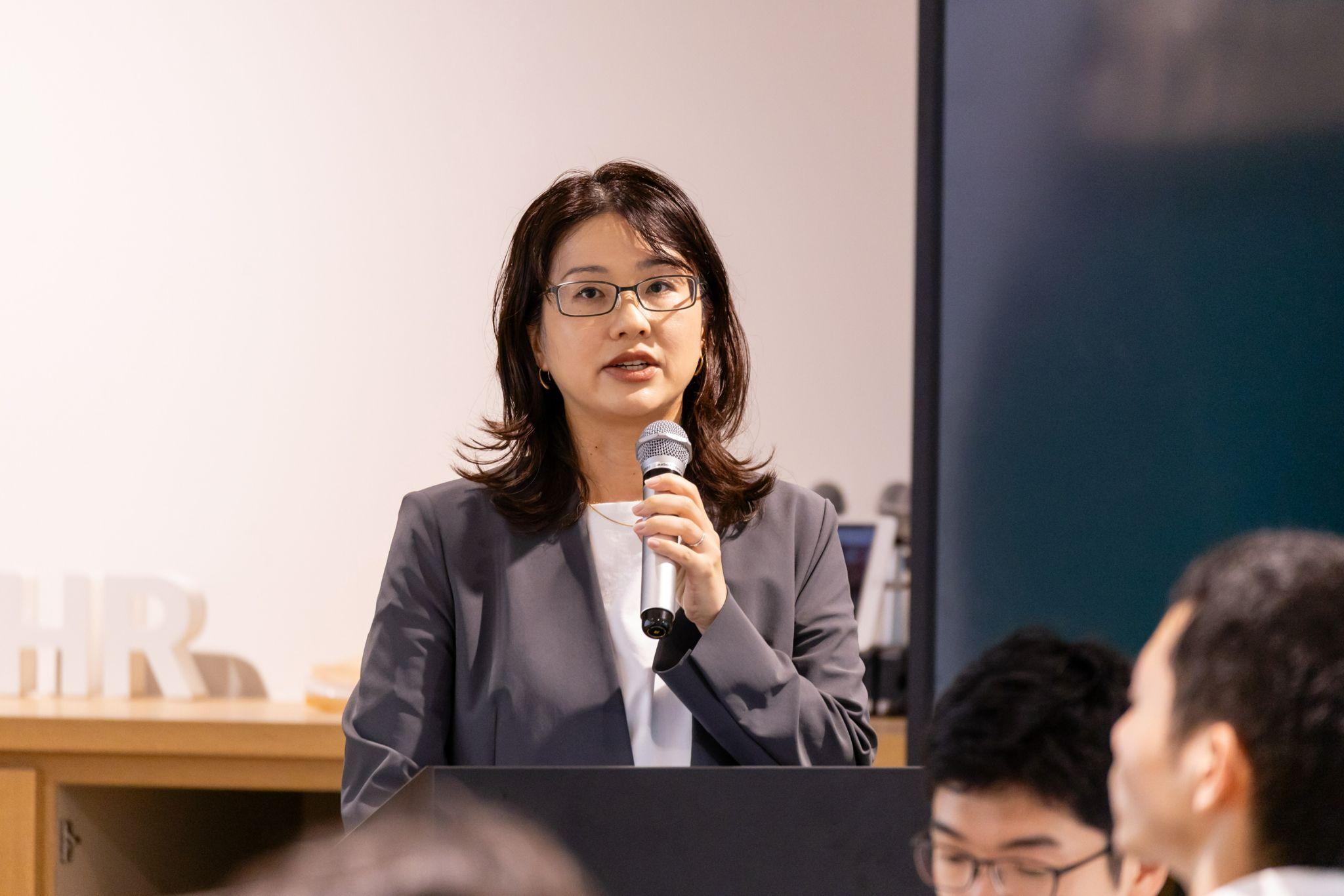 演台に立ち、聴衆に向かってマイクで話をしている眼鏡をかけたグレースーツの松栄さん 