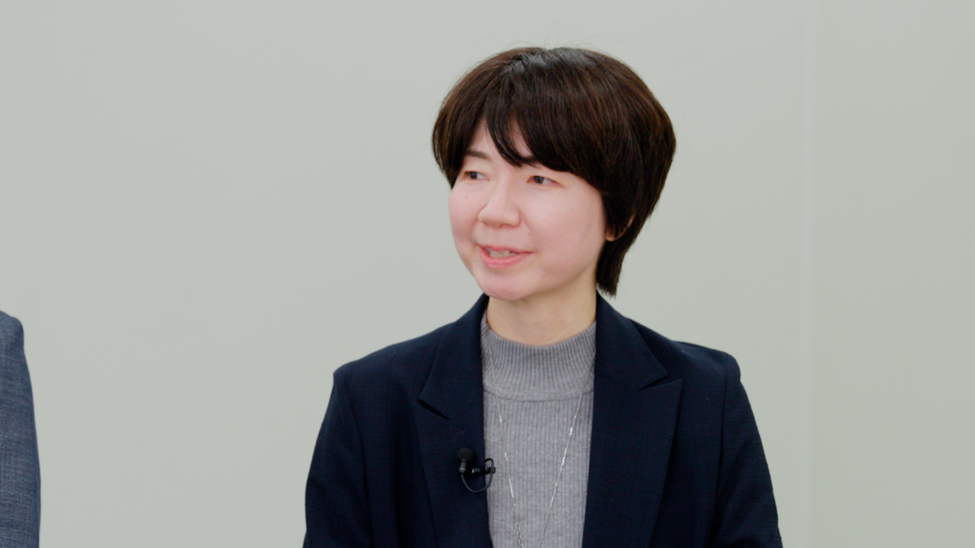 紺色のジャケットを着用し、左側を向いて穏やかな表情で話すショートヘアの女性。