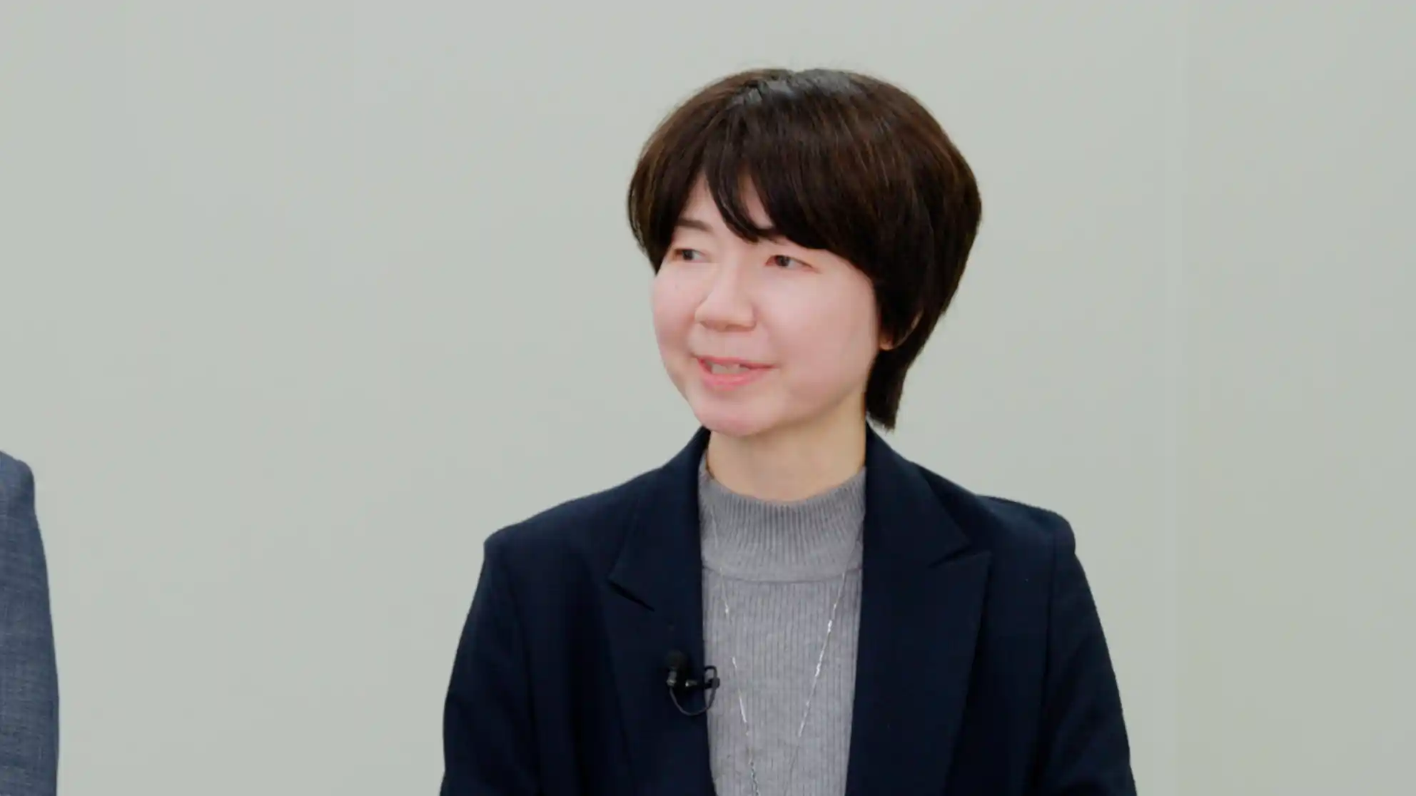 紺色のジャケットを着用し、左側を向いて穏やかな表情で話すショートヘアの女性。