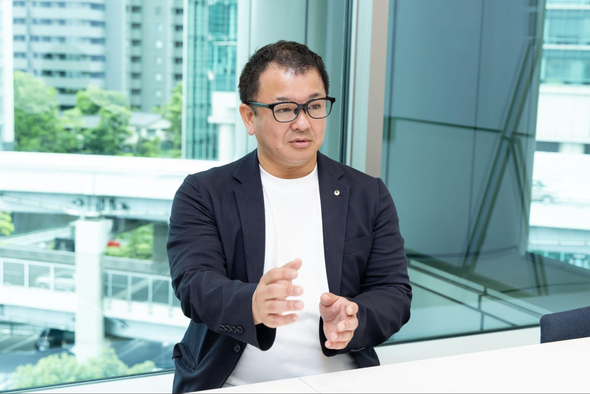 オフィスの窓際で、身振り手振りを交えてインタビューに答える田川さん
