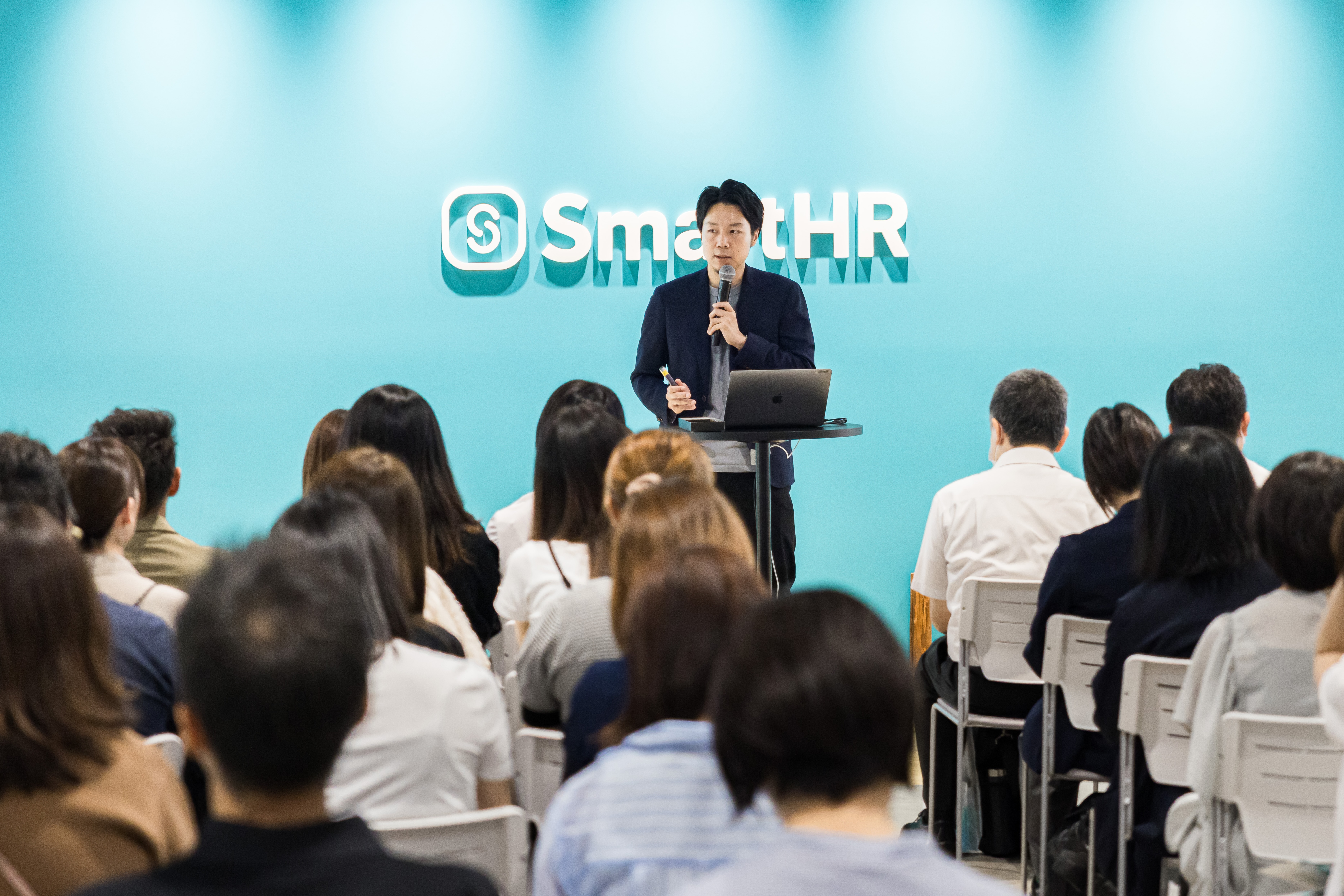 「SmartHR」のロゴが掲げられた青い壁を背景に、マイクを持って参加者に向けて話す芹澤