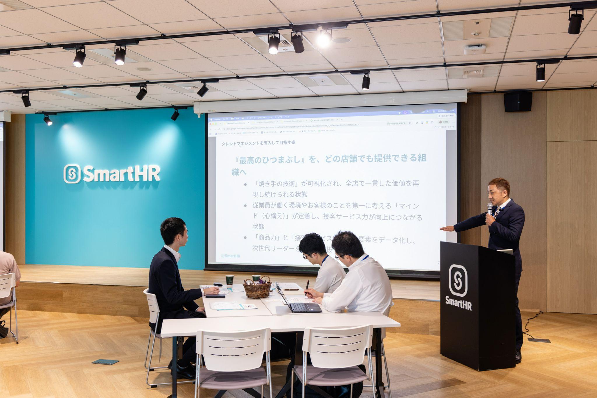 SmartHRロゴ入りの会場でのプレゼンテーション。「『最高のひつまぶし』を、どの店舗でも提供できる組織へ」というスクリーンの前で月井さんが話し、聴衆がテーブルでメモを取る様子。