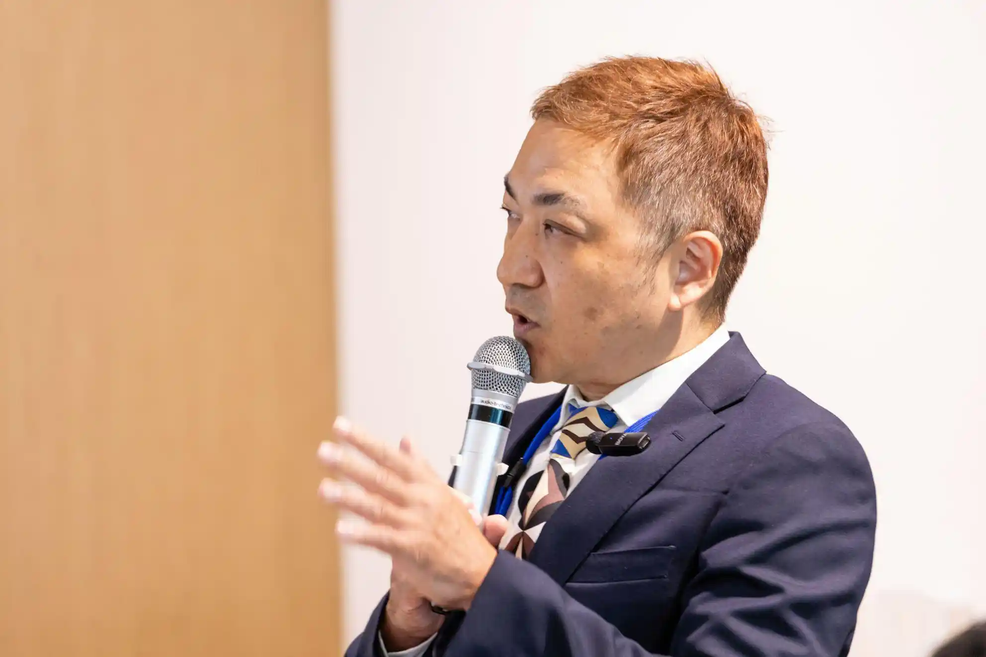 マイクを握り、手でジェスチャーを交えながら話す紺色のスーツを着た月井さん