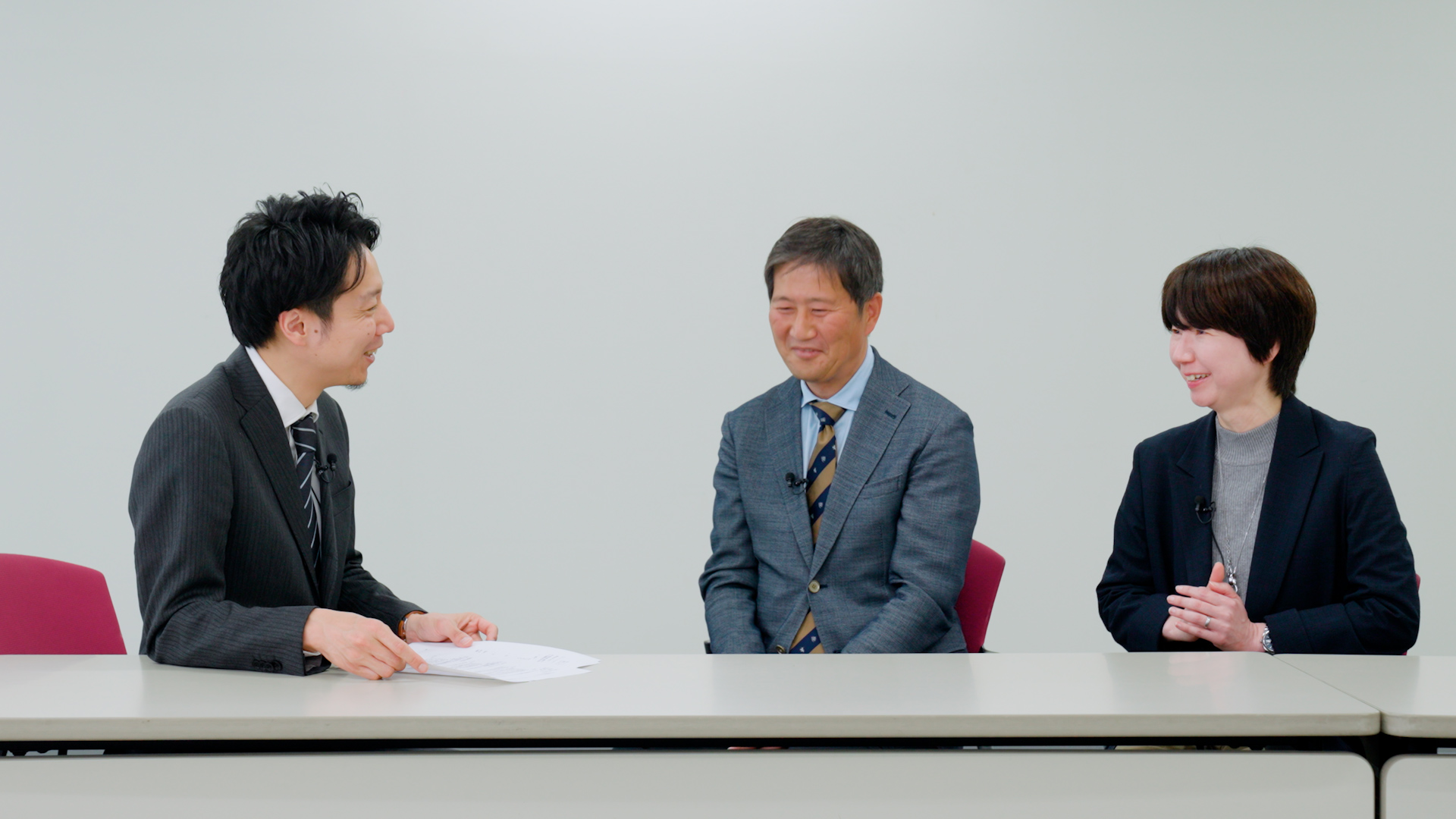 長机を挟んでスーツ姿のビジネスパーソン3名が笑顔で対談している様子。