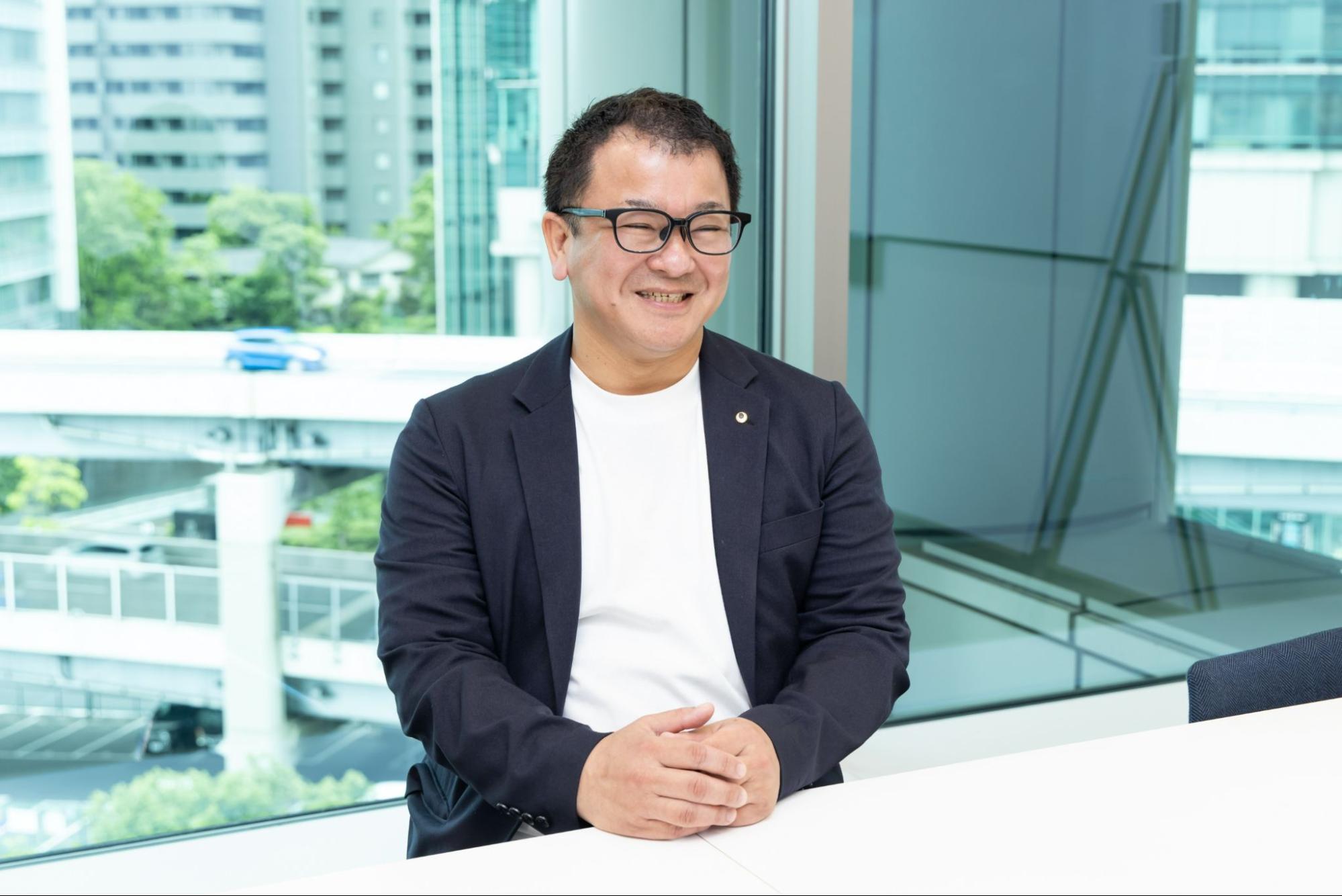 オフィスの窓際で白いテーブルに手を組み、穏やかな笑顔を見せる田川さん