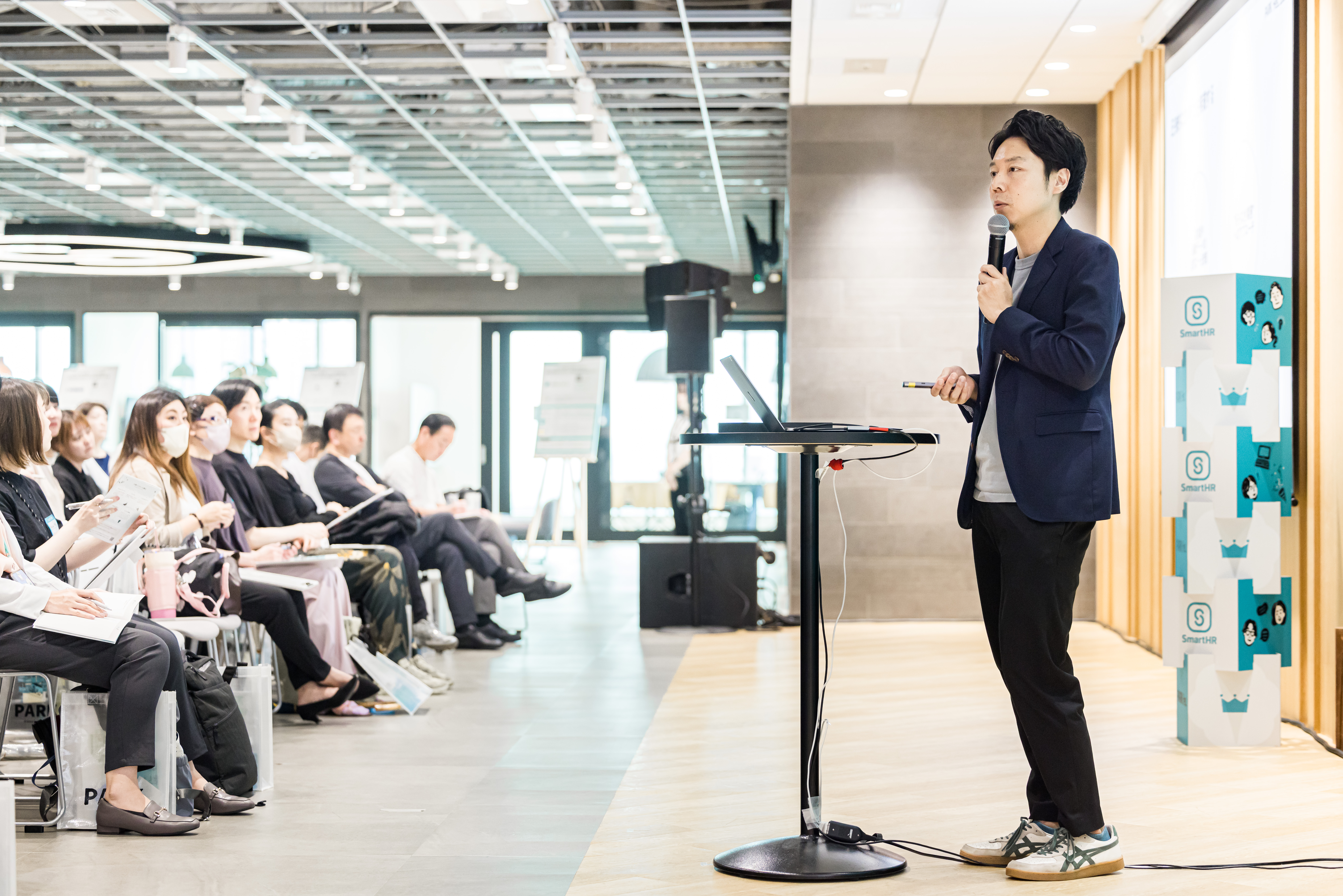 SmartHRのロゴが置かれたセミナー会場で、マイクを持って登壇する芹澤と着席して話を聴く参加者