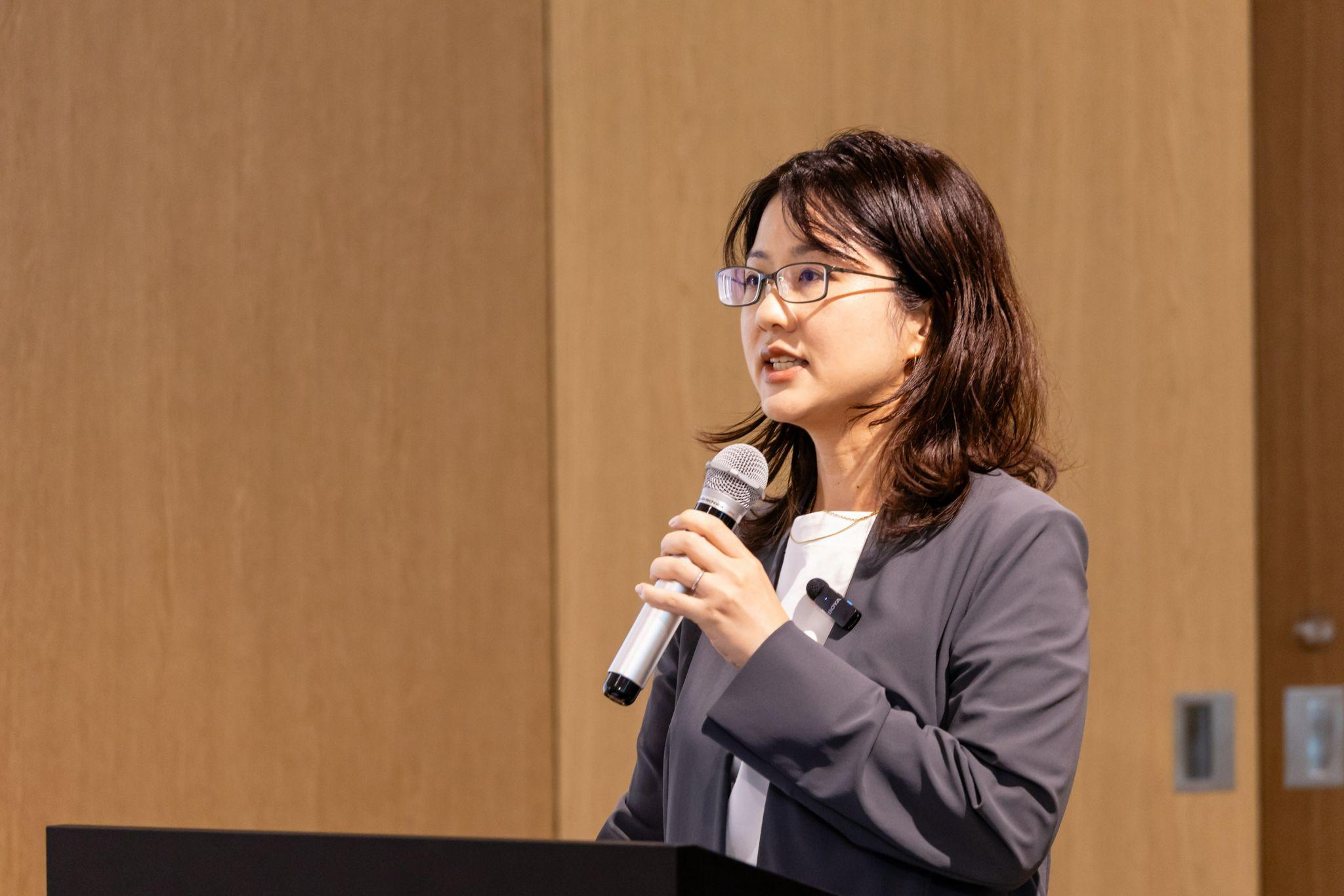 演台に立ち、マイクを持って話をしている眼鏡をかけたグレースーツの松栄さん 