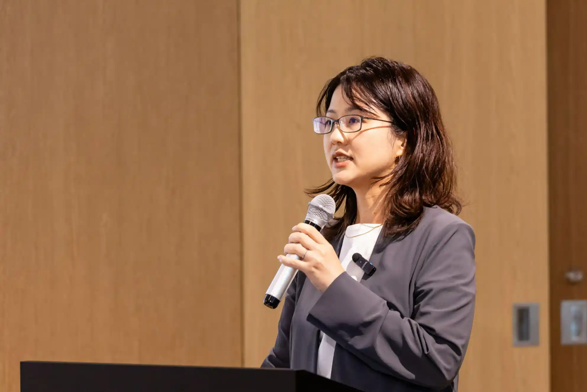 演台に立ち、マイクを持って話をしている眼鏡をかけたグレースーツの松栄さん