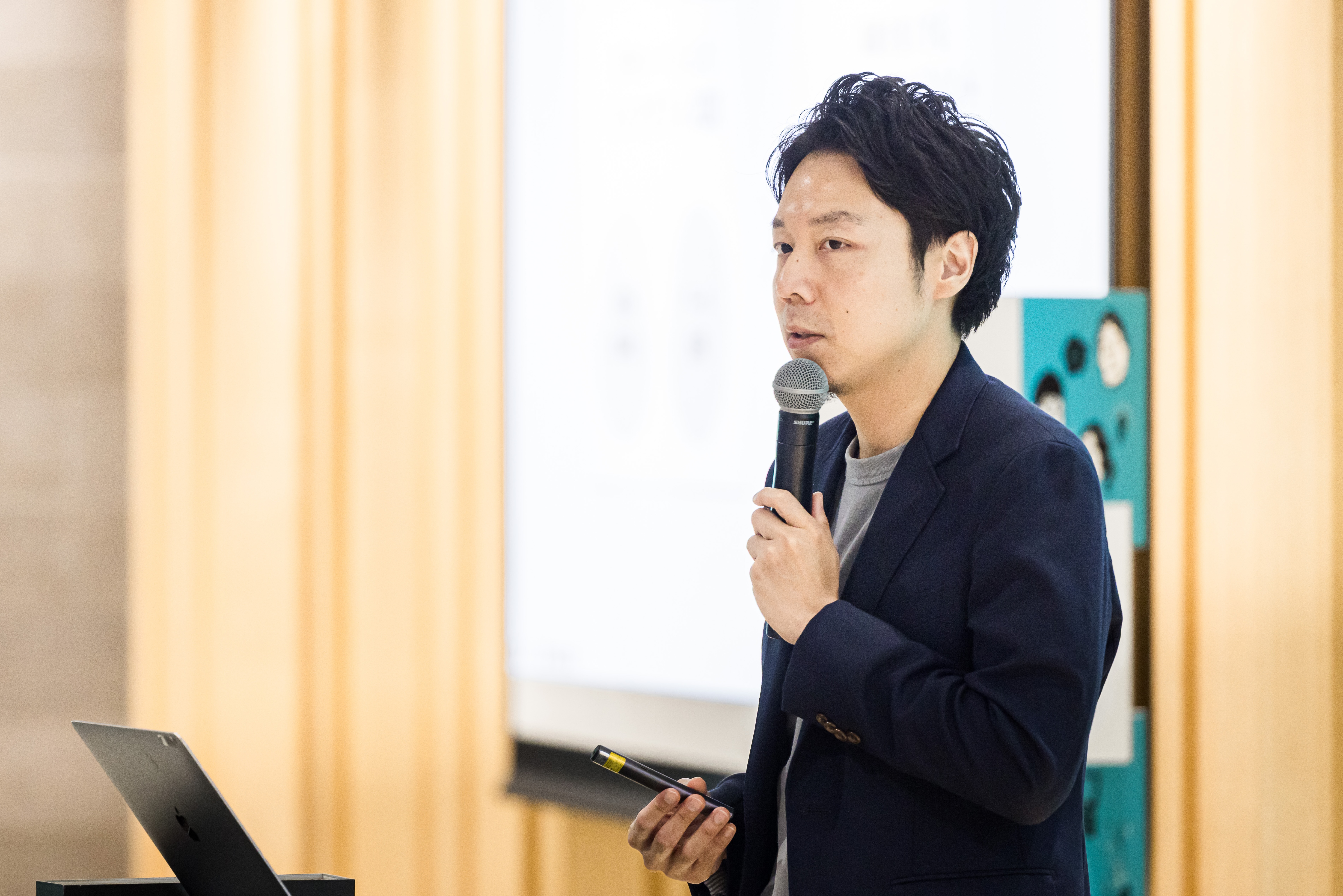 スクリーンを背に、マイクとポインターを持ってプレゼンテーションを行う芹澤