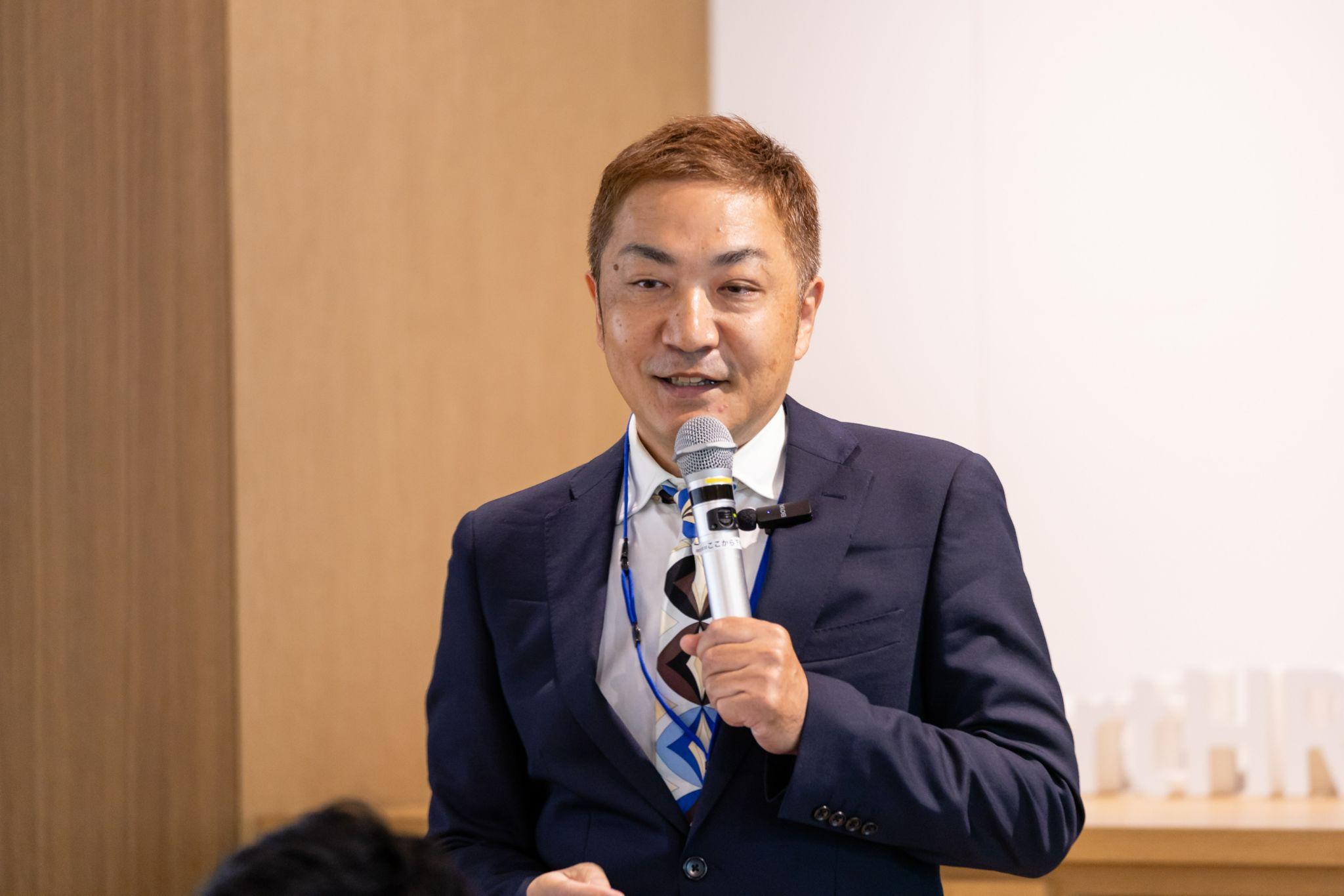 マイクを右手に持ち、スピーチをしている紺色のスーツを着た月井さん