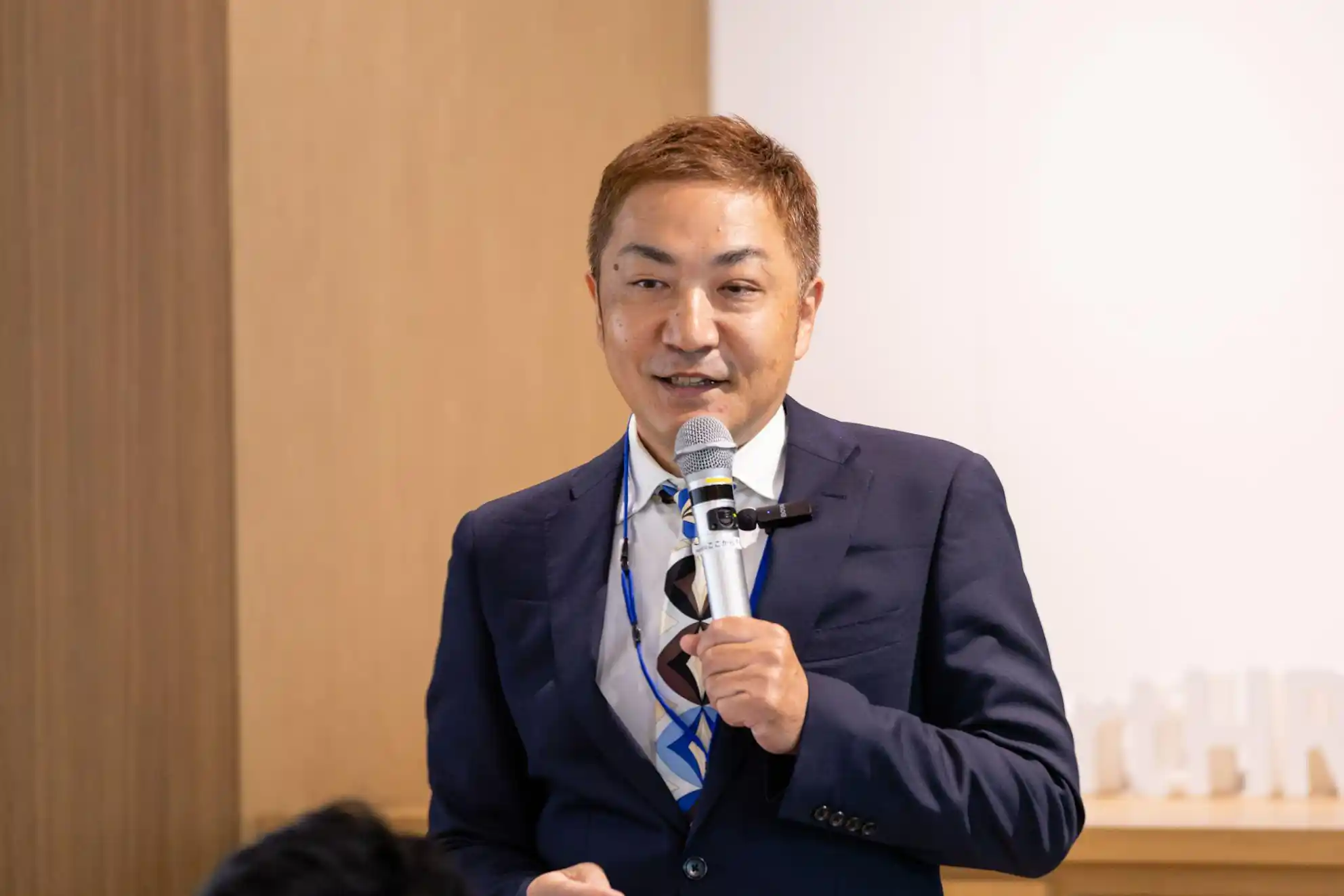 マイクを右手に持ち、スピーチをしている紺色のスーツを着た月井さん
