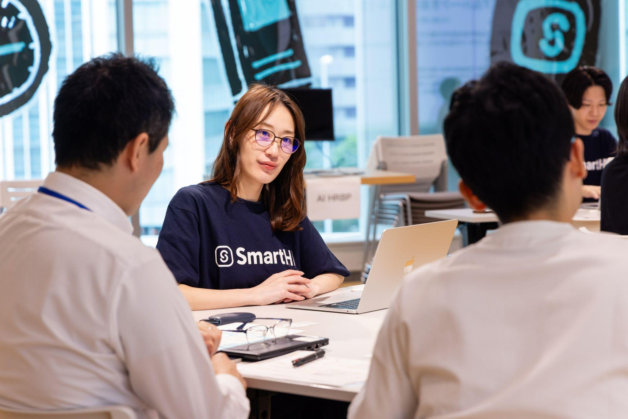 オフィスのテーブルでノートパソコンを開き、向かいに座る2人と話をしているSmartHRのTシャツを着た女性