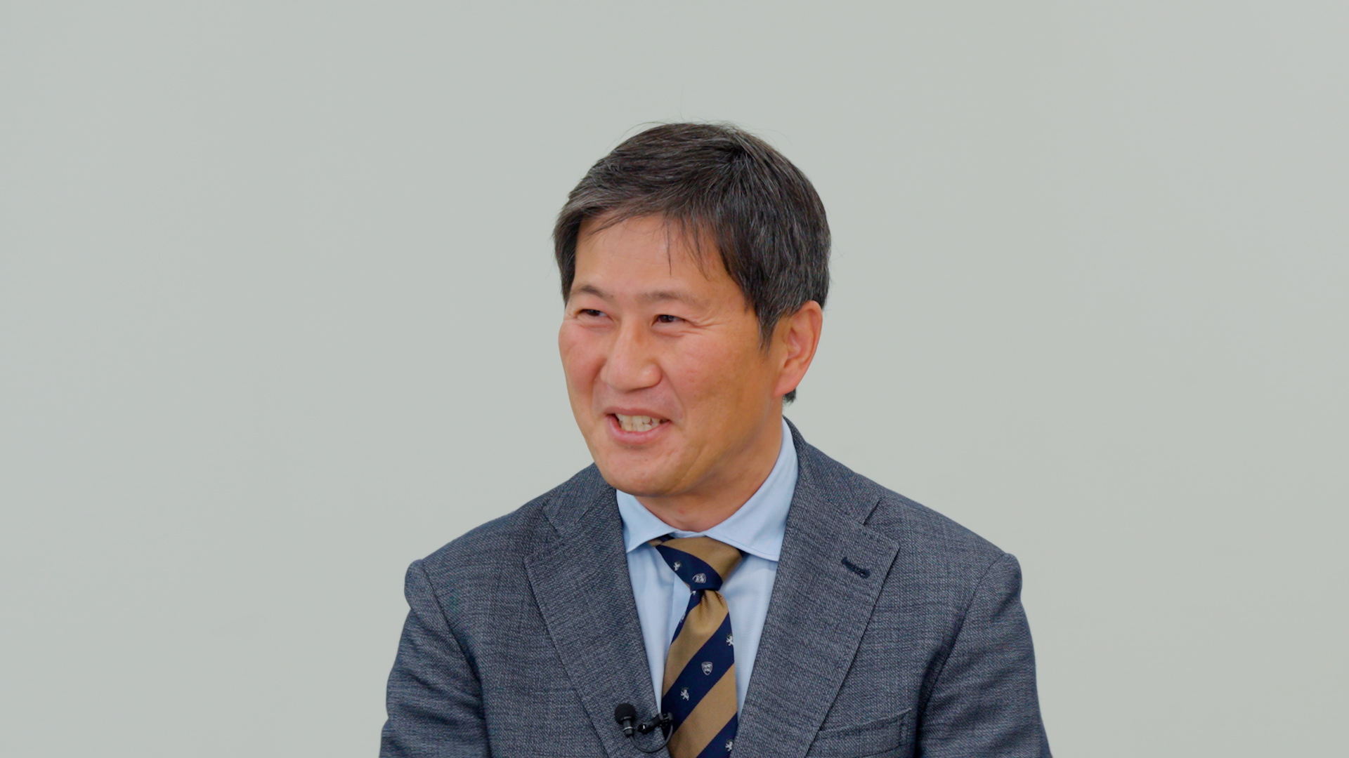 グレーのジャケットとネクタイを着用し、左側を向いて笑顔で話す男性。