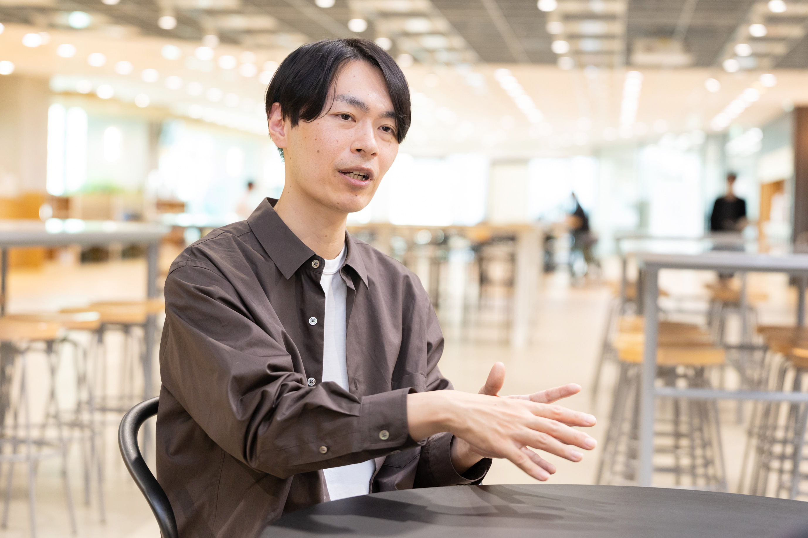 開放的なオフィスで、テーブルに座り手を使って説明をしている茶色いシャツの男性