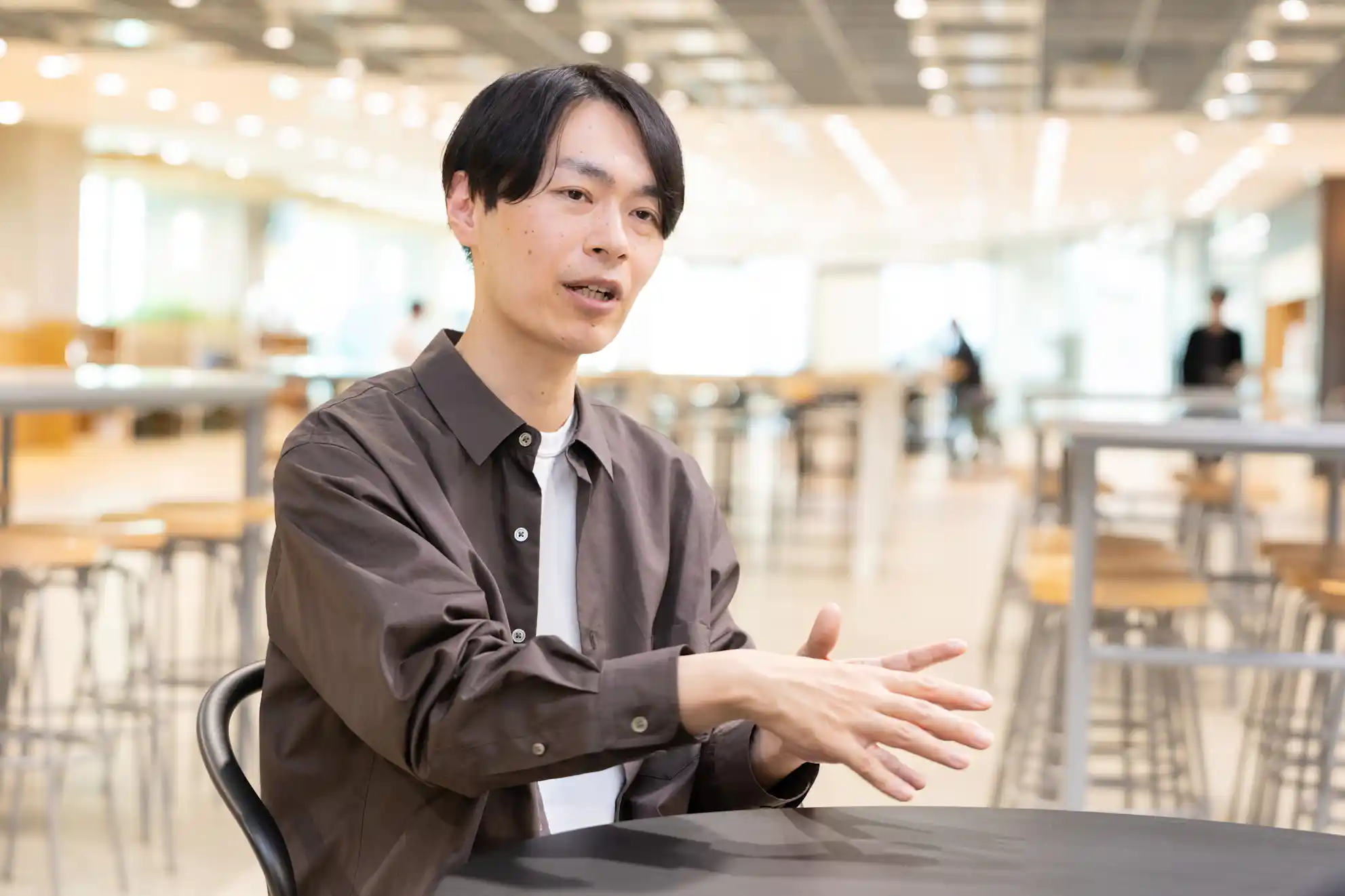 開放的なオフィスで、テーブルに座り手を使って説明をしている茶色いシャツの男性