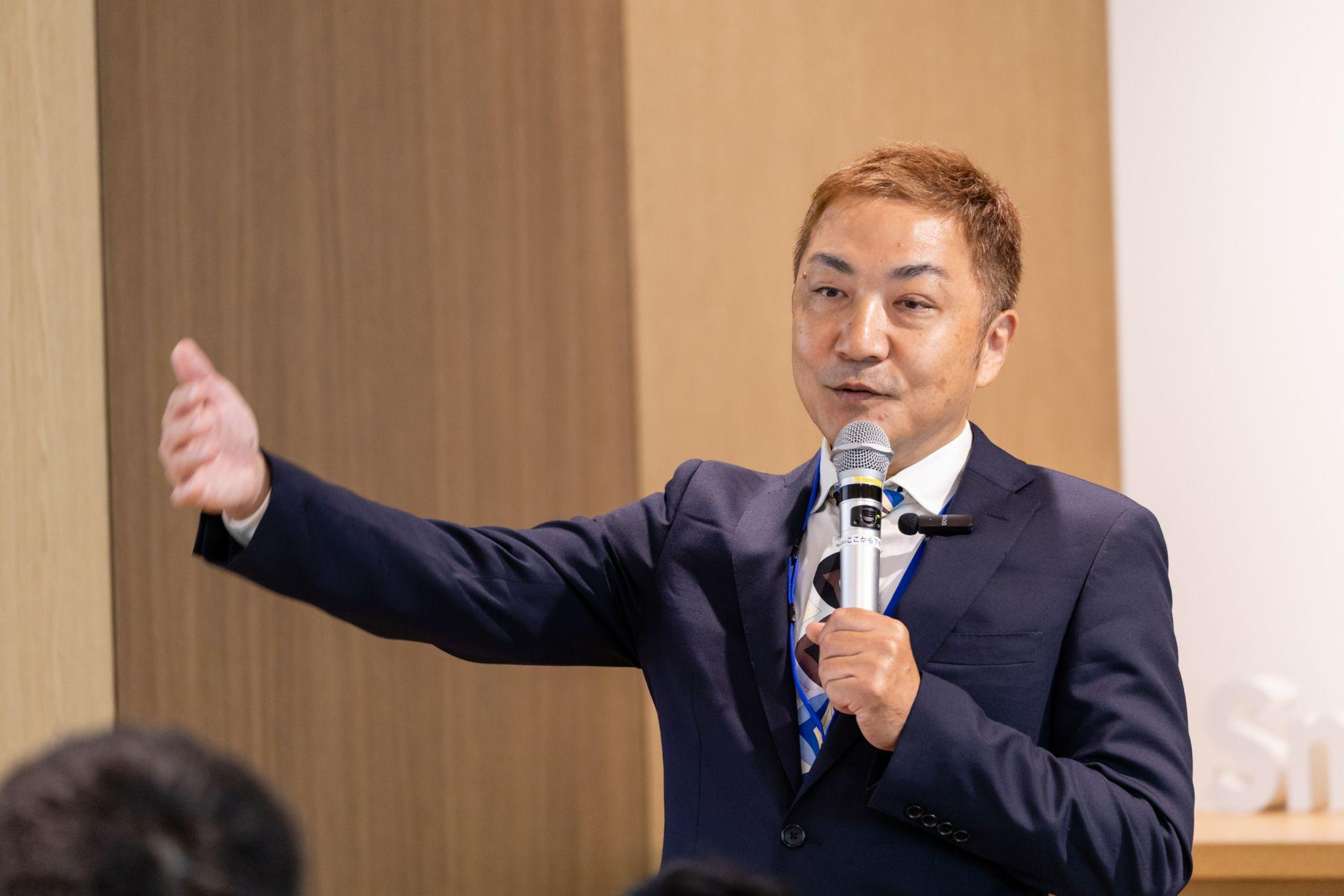 マイクを持ち、左手を広げてスピーチをする紺色のスーツを着た月井さん