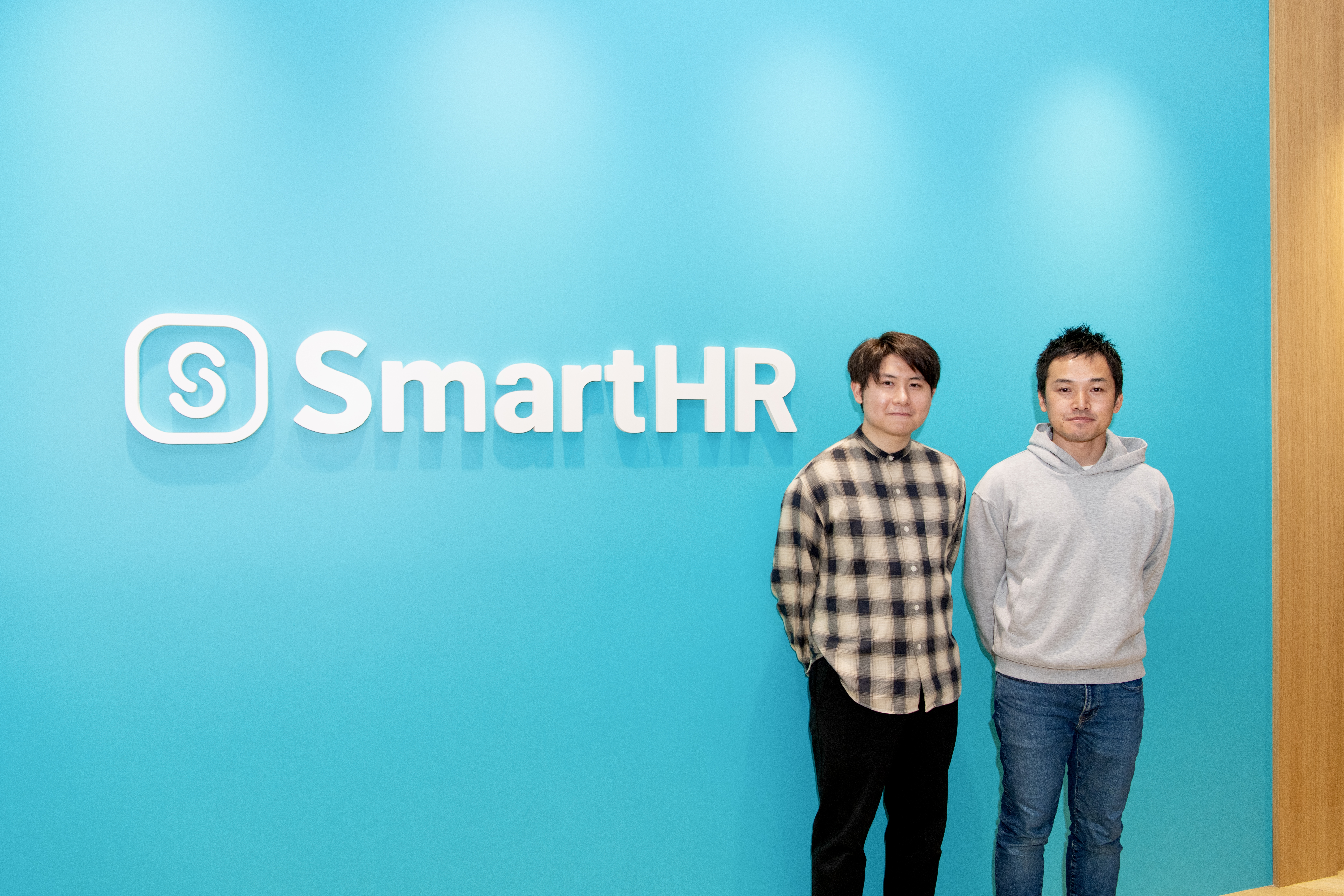 SmartHRのロゴの前で、今後の展望に向けて意気込みを見せる向高と谷さん