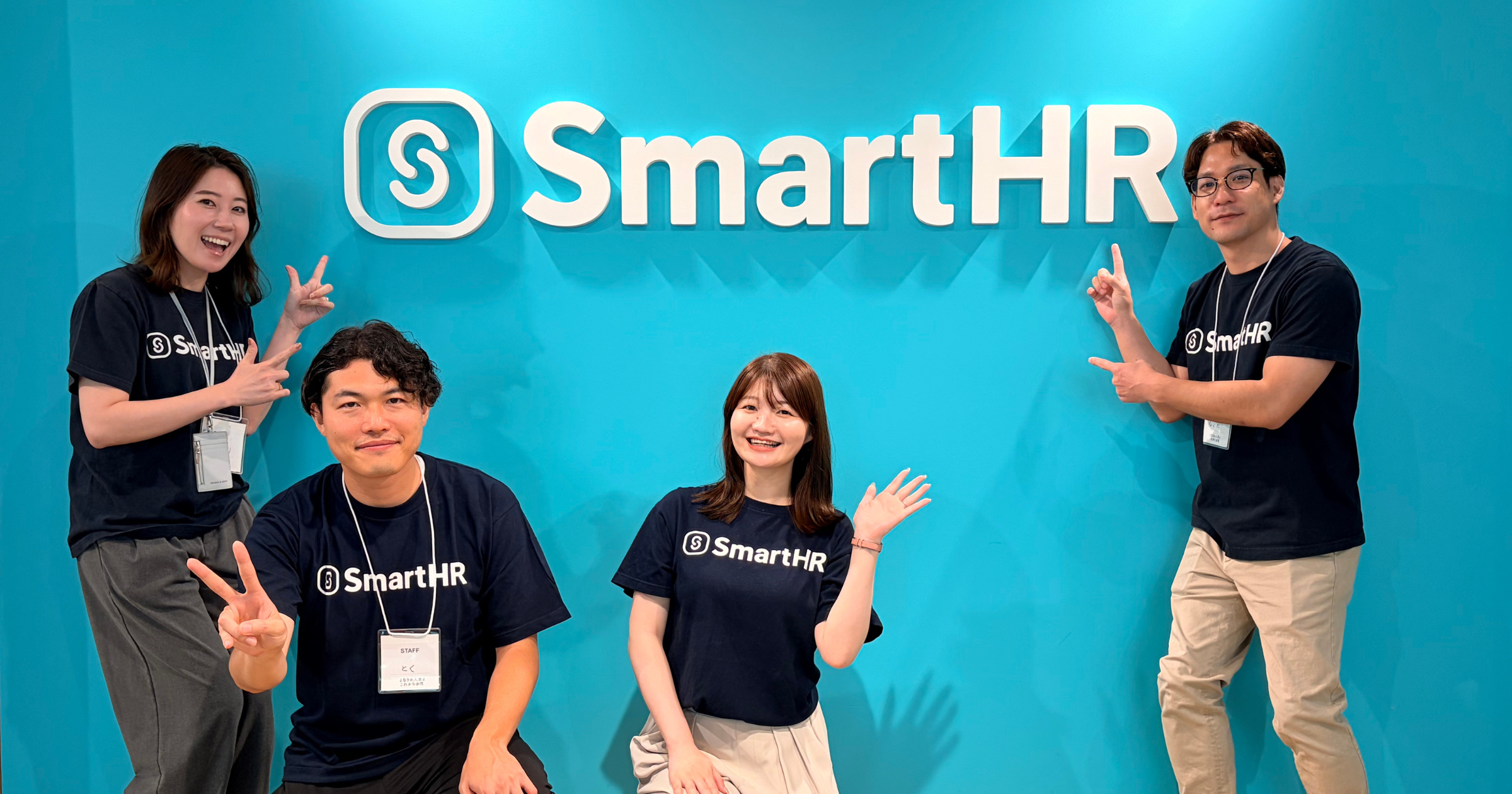水色の壁に掲げられたSmartHRのロゴの前で、お揃いのロゴ入りTシャツを着て笑顔でポーズをとる4人のスタッフ