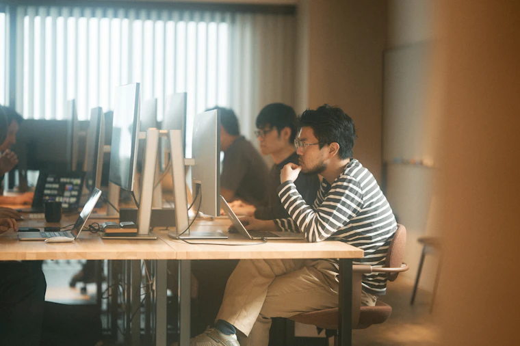 オフィス内。大型モニターとノートPCを前に、顎に手を当てて画面を見つめる塚田と、奥に並ぶ岩本。
