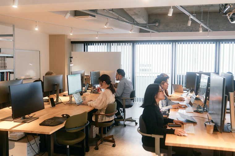 オフィス内。対面式の長机にモニターやPCが並び、複数の人が椅子に座って作業をしている。
