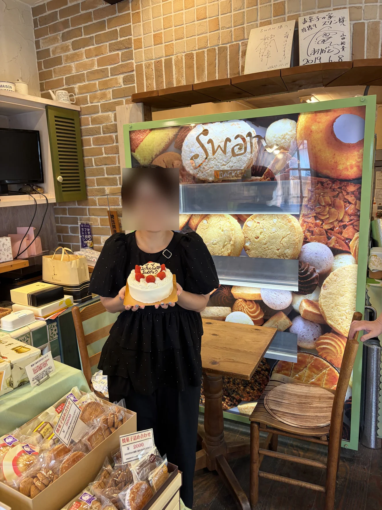 店内の焼き菓子パネルの前でホールケーキを両手で持つ妻