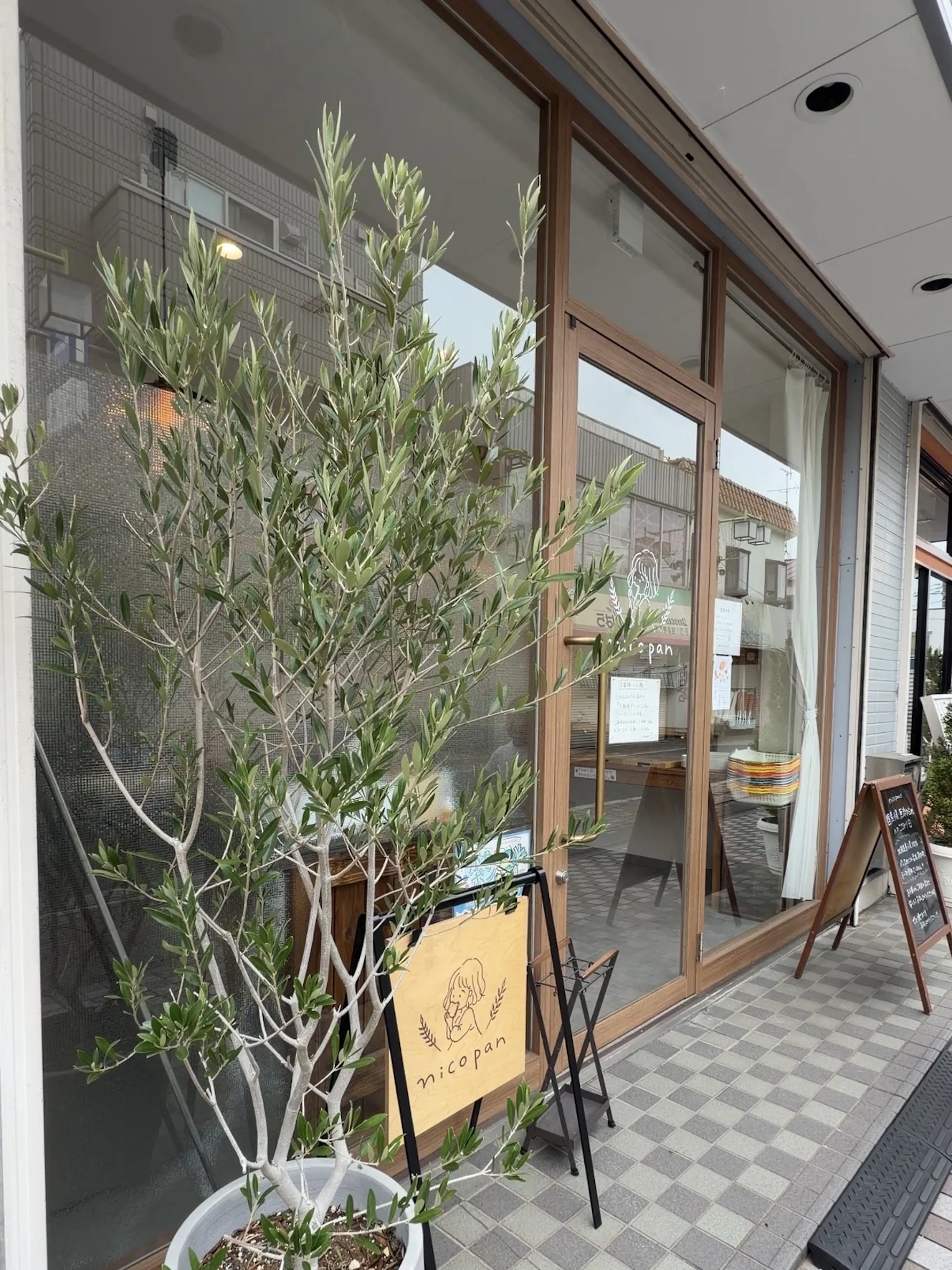 nicopanの外観。大きなオリーブの鉢植えと、麦の穂とパンを持つ女性のロゴが入った木製のA型看板