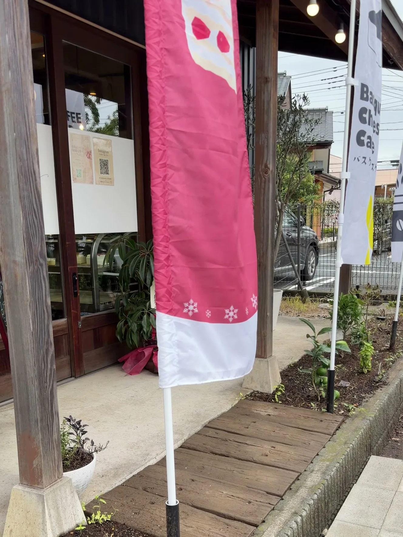 木の柱の店先に立つピンクののぼりとBasque Cheese Cakeののぼり。奥に濃い茶色の木枠のドア