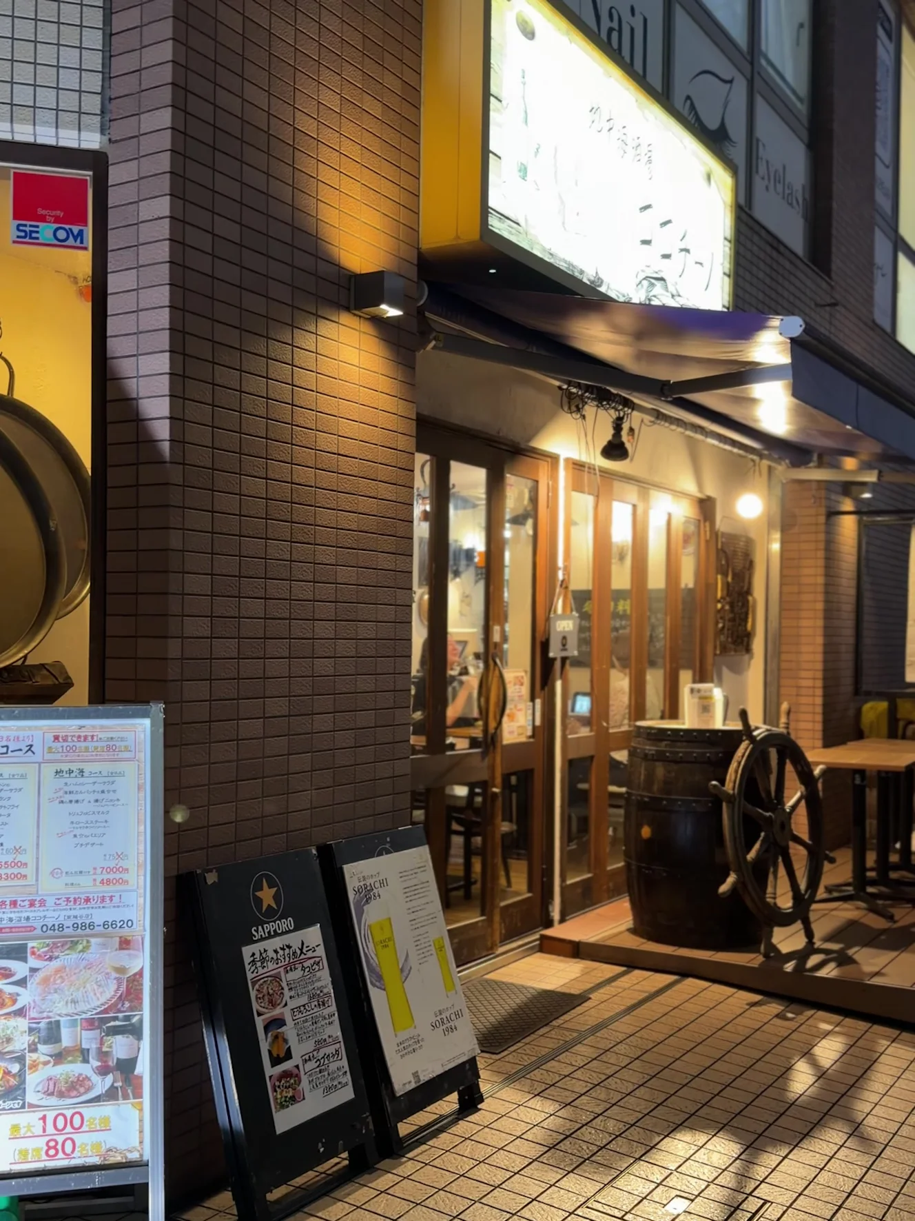 地中海酒場ココチーノ 南越谷店の夜の外観。黄色い看板の下に樽と舵輪、入口の立て看板がある