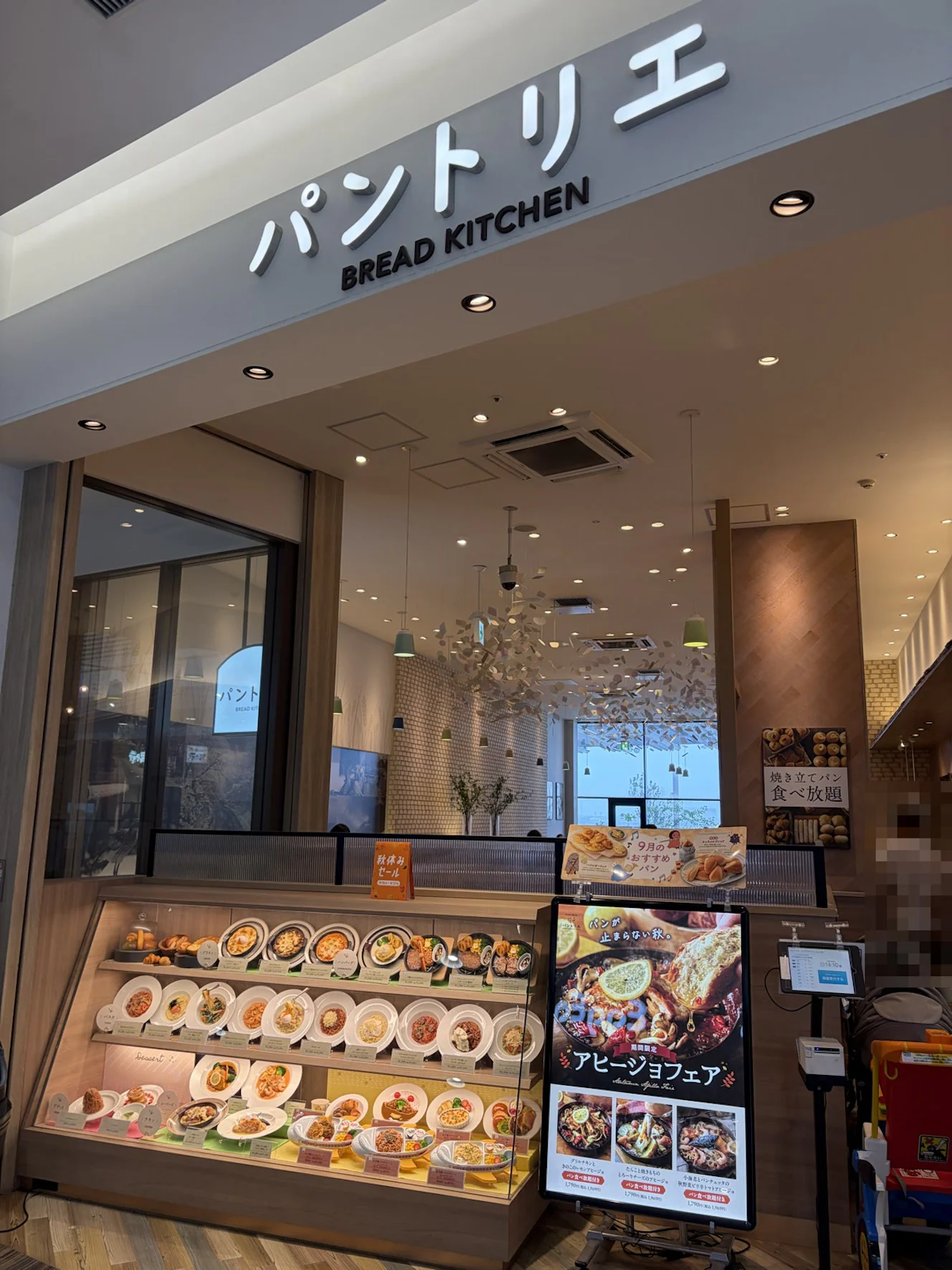 パントリエ レイクタウンkaze店の外観。店頭に食品サンプルのショーケースとアヒージョフェアのポスターがあり、右側に発券機が置かれている