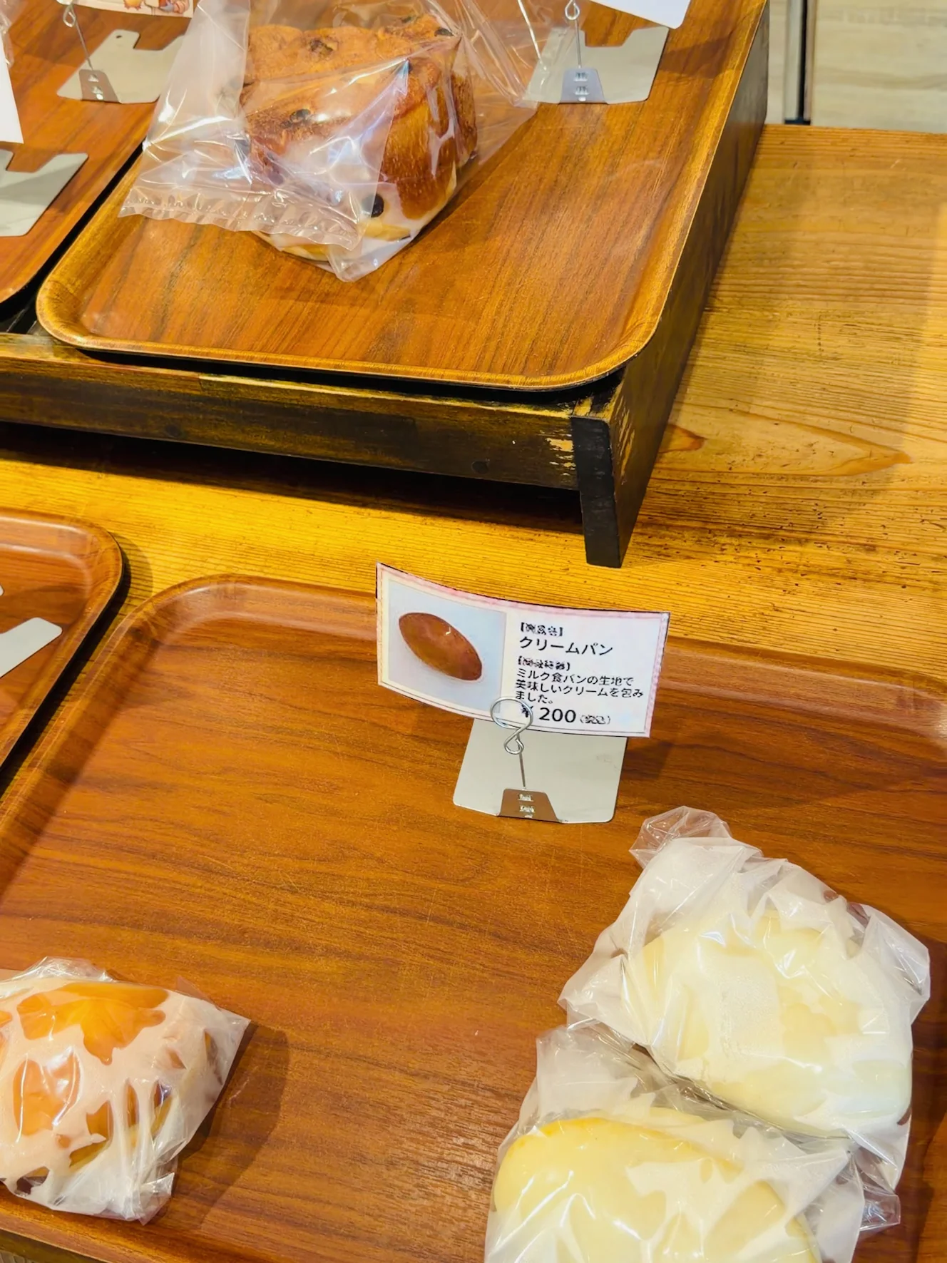 木のトレーに並んだ袋入りのパンと値札。クリームパン200円の札が立っている