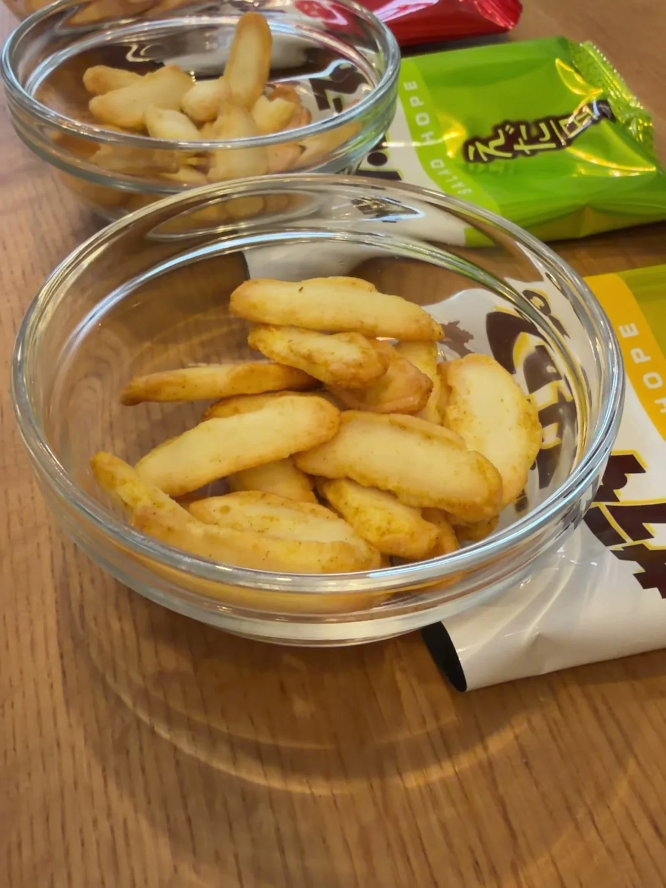 ガラスの器に出したカレー味のあられ。黄色いカレー味の小袋の上に器が乗り、奥に緑のえだ豆味の小袋