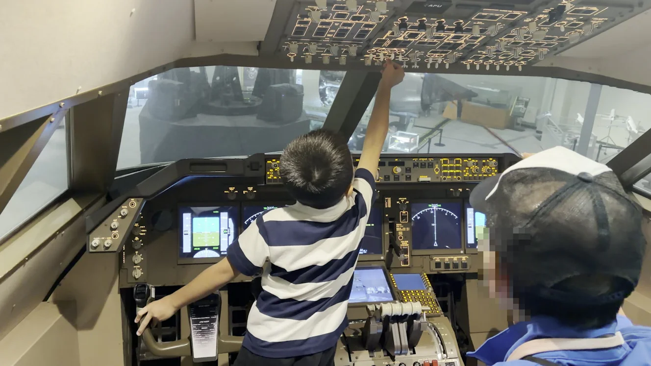 ボーイング747-400のコクピット内。操縦席で頭上のパネルに手を伸ばす子どもの後ろ姿と、横にいる青いジャンパーの係員。窓の向こうに大型模型の台座が見える