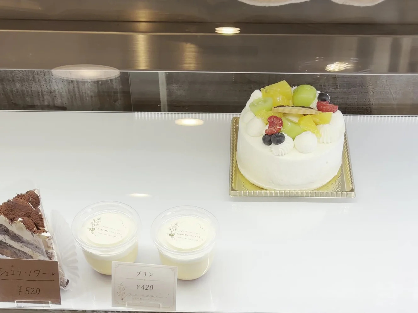 ショーケースのフルーツを乗せた白いホールケーキ。手前にプリン420円とショコラ・ノワール520円の札