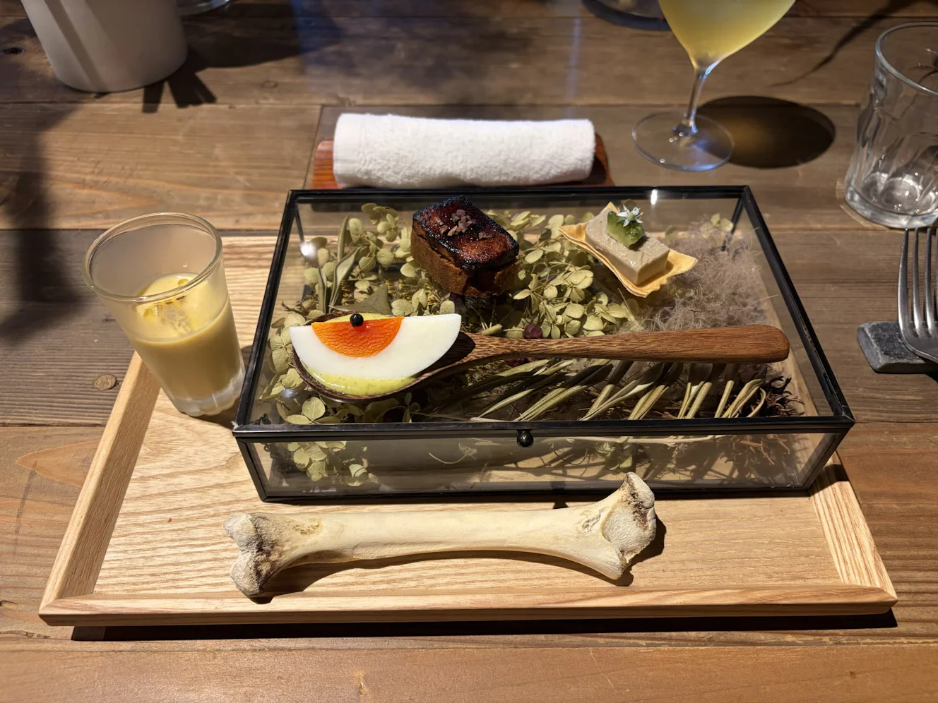 木のトレーに載った前菜。飾りを入れたガラスの箱の上に小さな料理、横にコーンスープのグラス、手前に骨の飾り