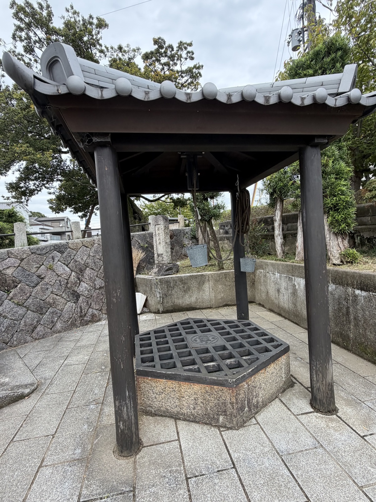 瓦屋根に覆われた井戸。六角形の石の井筒に格子状の鉄蓋がかぶせられ、柱に釣瓶の縄と2つのバケツが下がる