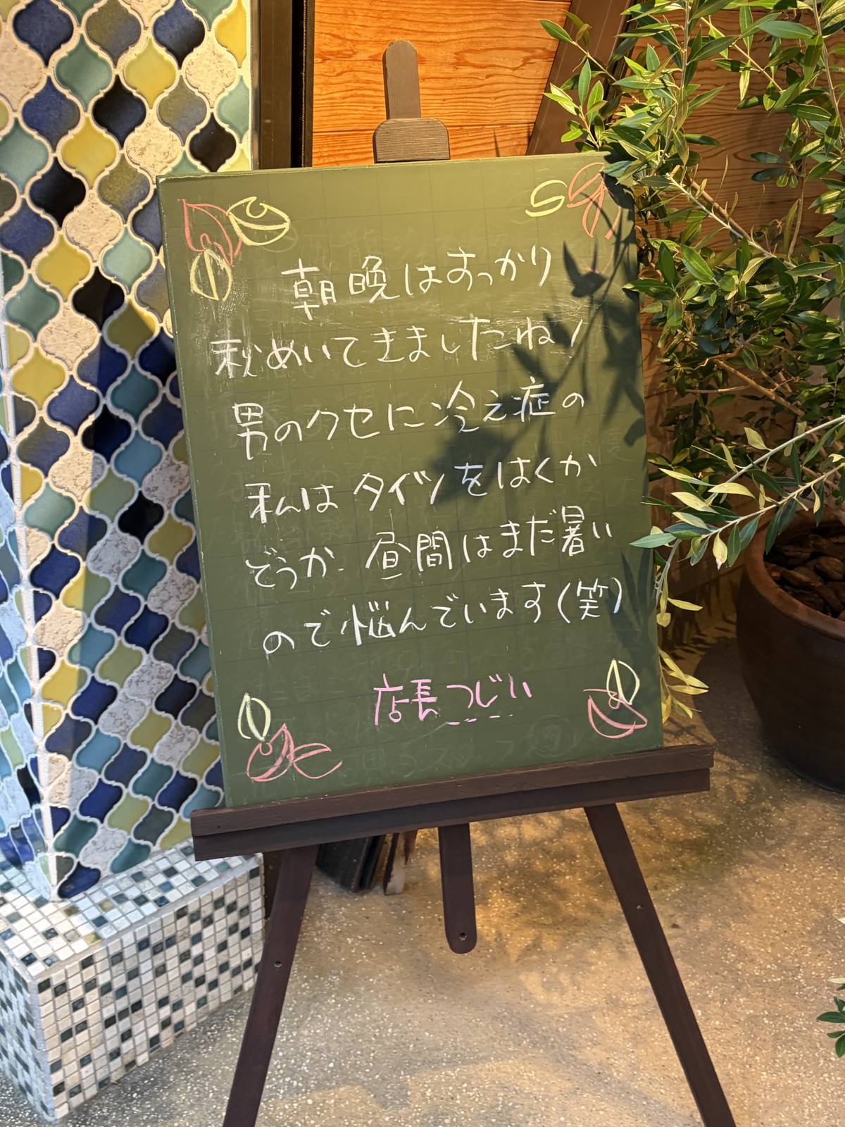 緑色の黒板に白いチョークの手書き。「朝晩はすっかり秋めいてきましたね！男のクセに冷え症の私は…」と、ピンクで「店長つじい」