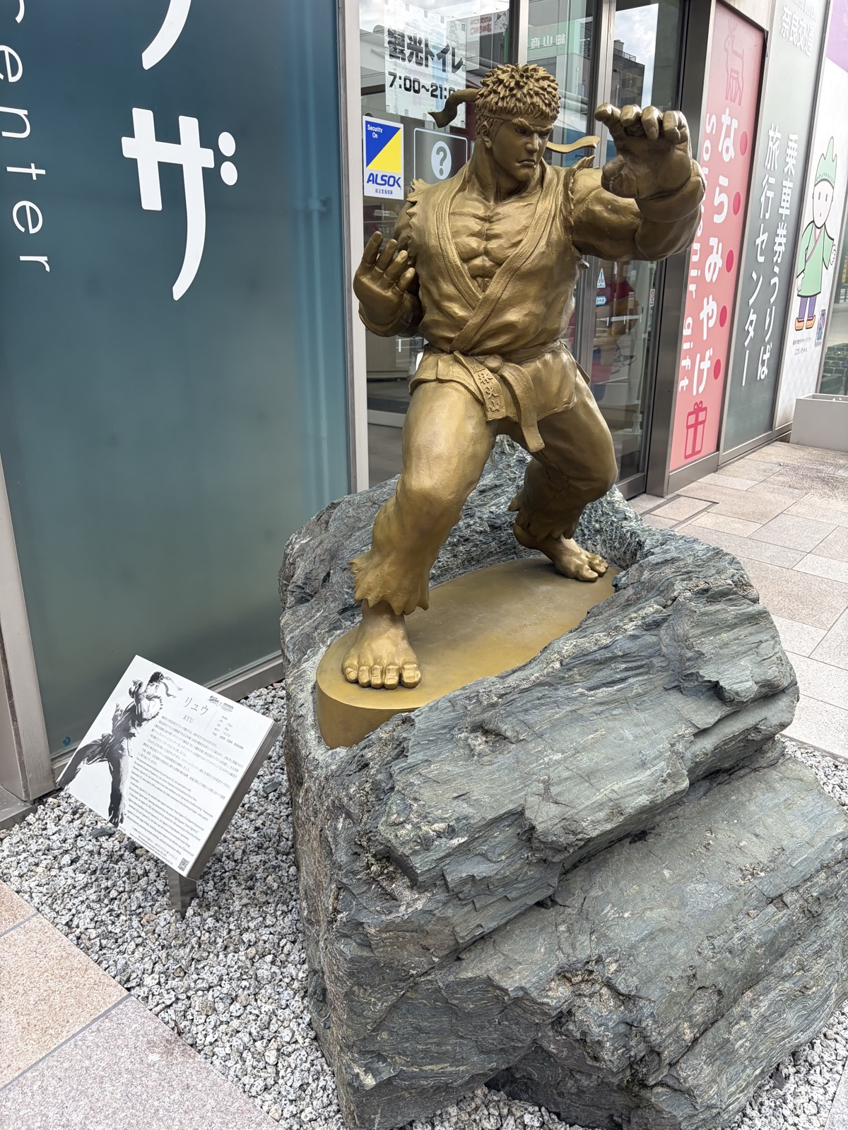 岩の台座の上で構える金色のリュウの立像。左手前に「リュウ」の解説板が立ち、背後は観光案内所のガラス扉