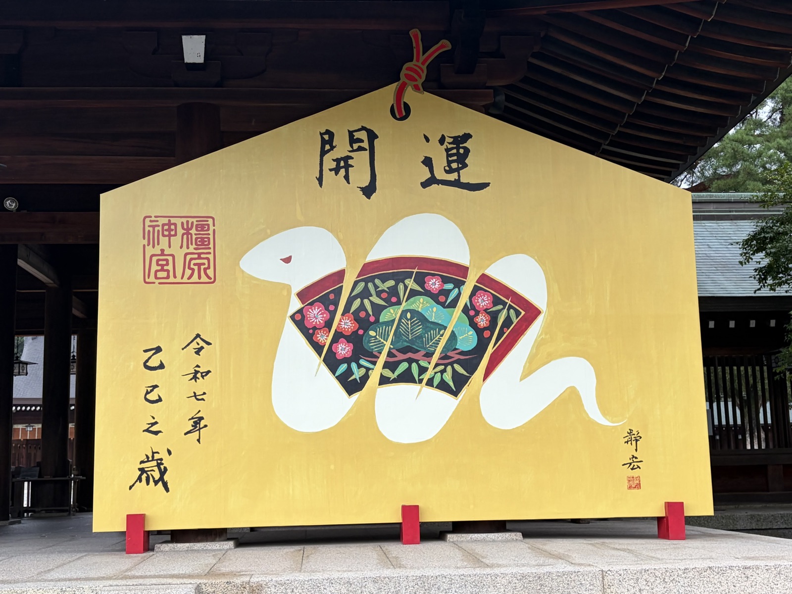 「開運」と大書された金色の巨大絵馬。扇の絵柄に白い蛇が巻きつき、左に「令和七年 乙巳之歳」の文字