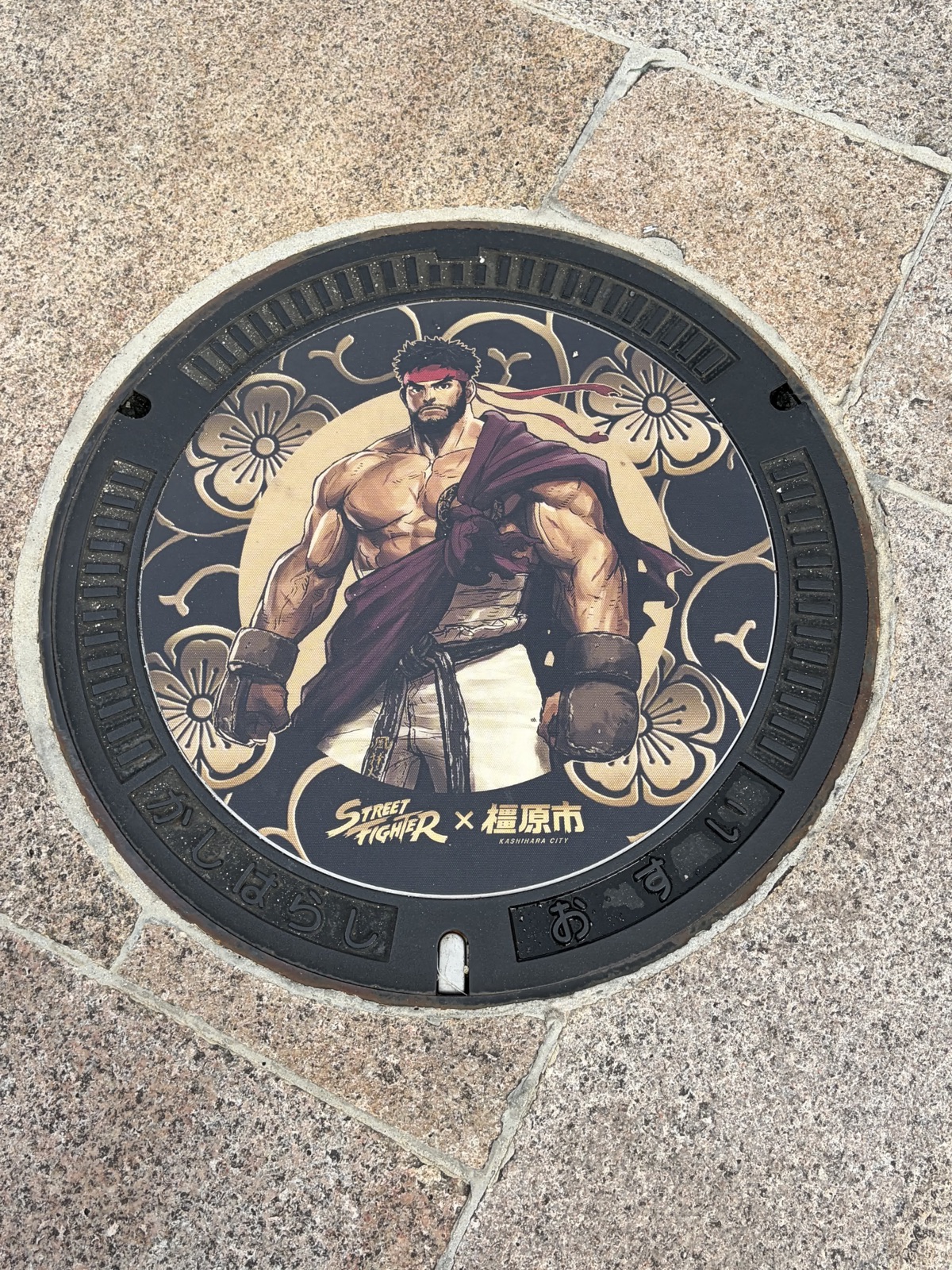 石畳にはめ込まれた黒い丸いマンホール蓋。鉢巻き姿のリュウが描かれ、下に「STREET FIGHTER × 橿原市」