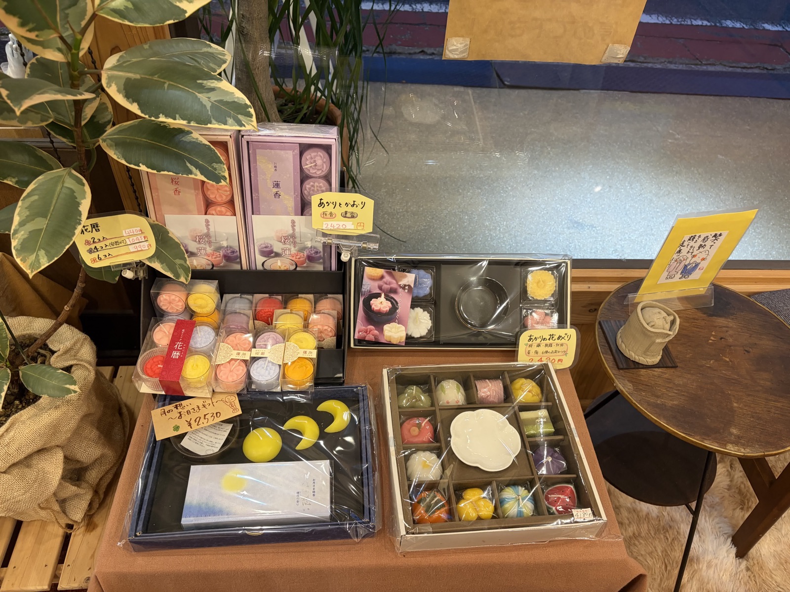 お香とキャンドルの並ぶ台。蓮や桜の花のかたちのキャンドル、月のかたちのもの、「あかりとかおり 2420円」などの手書き値札